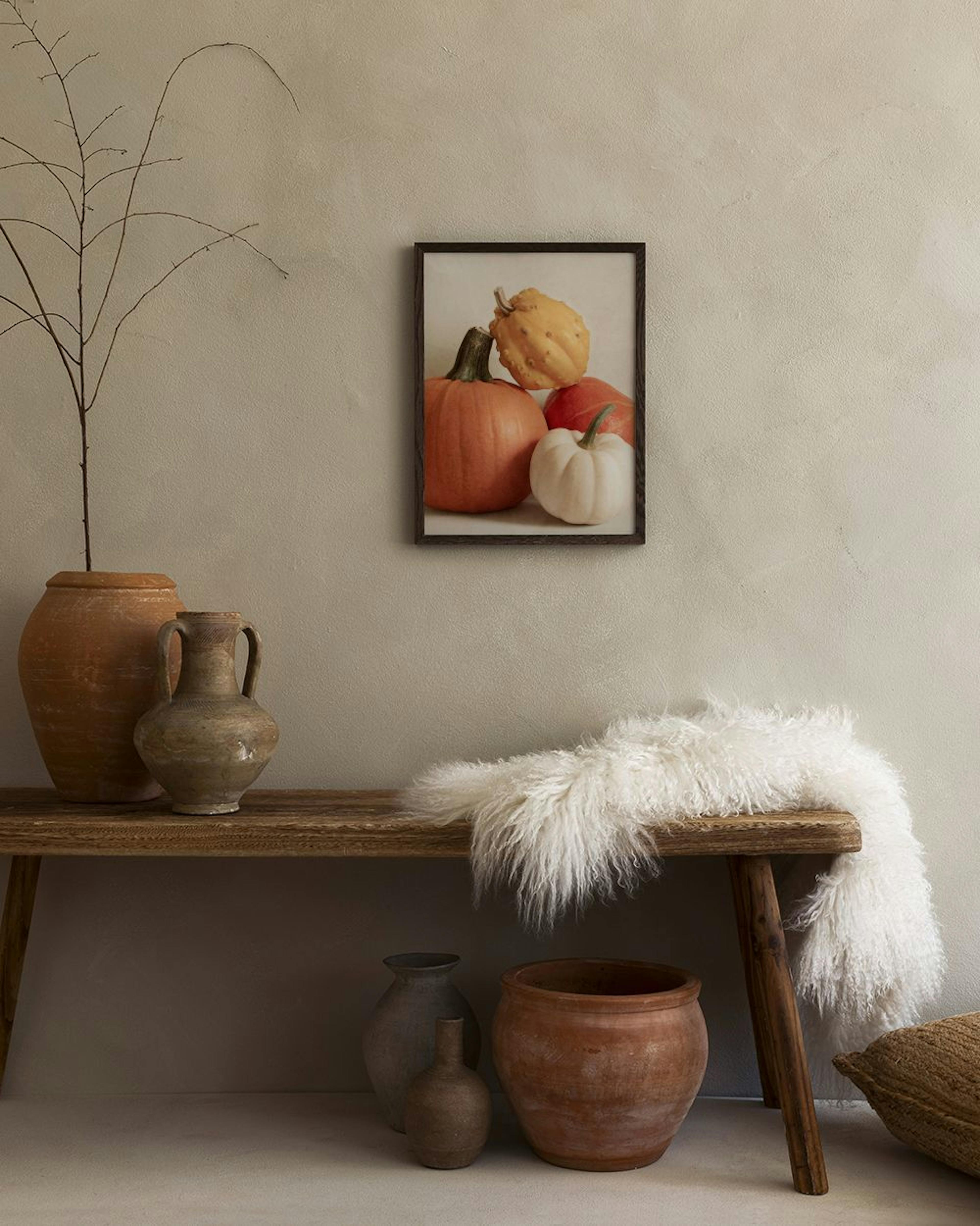 Poster van oranje, gele en witte pompoenen, hangend boven een houten bankje met aardewerken potten en een wit kleed.