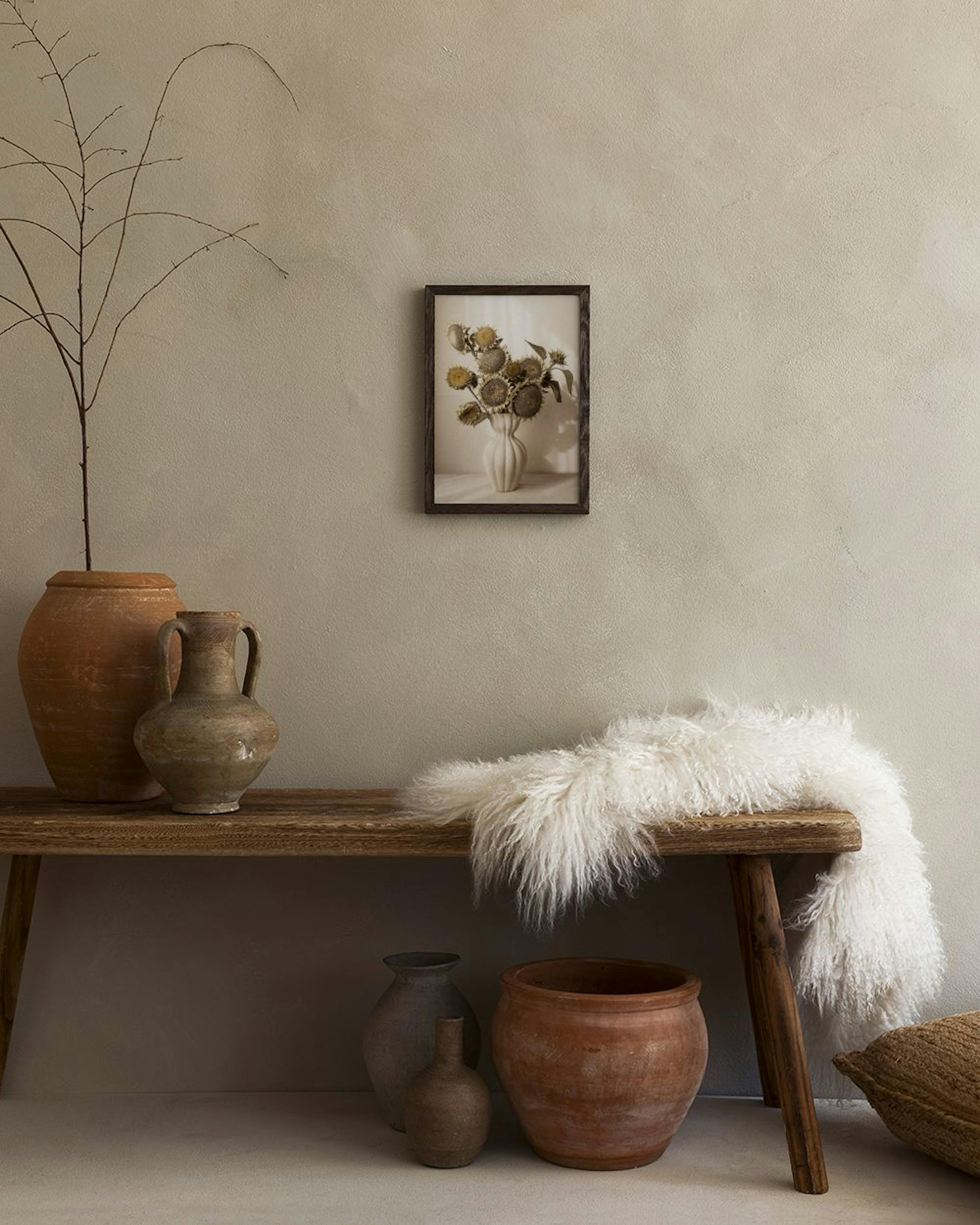Affiche de tournesols séchés dans un vase, accrochée au-dessus dun banc en bois avec des poteries et une fourrure blanche.