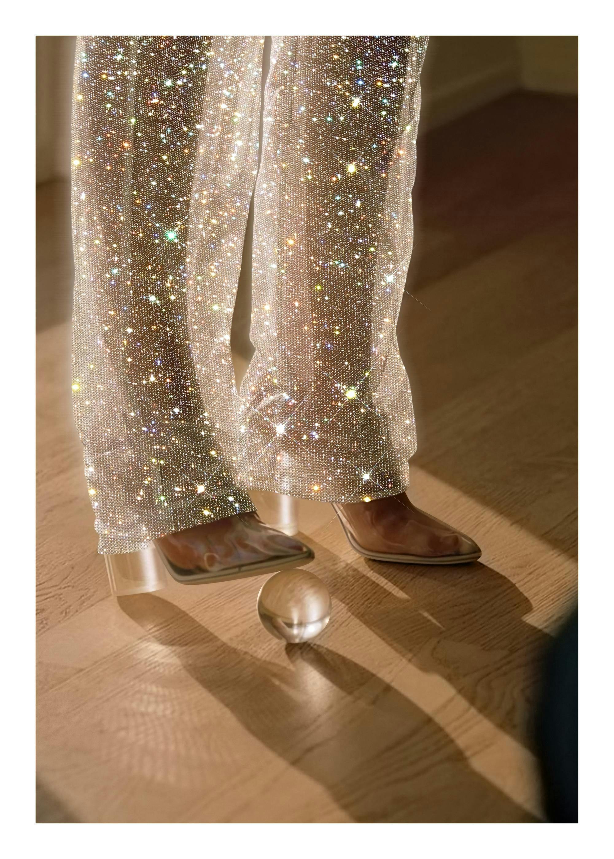 Photograph of sparkling trousers and shoes, with a glass ball under the heel, creating a glamorous fashion print.