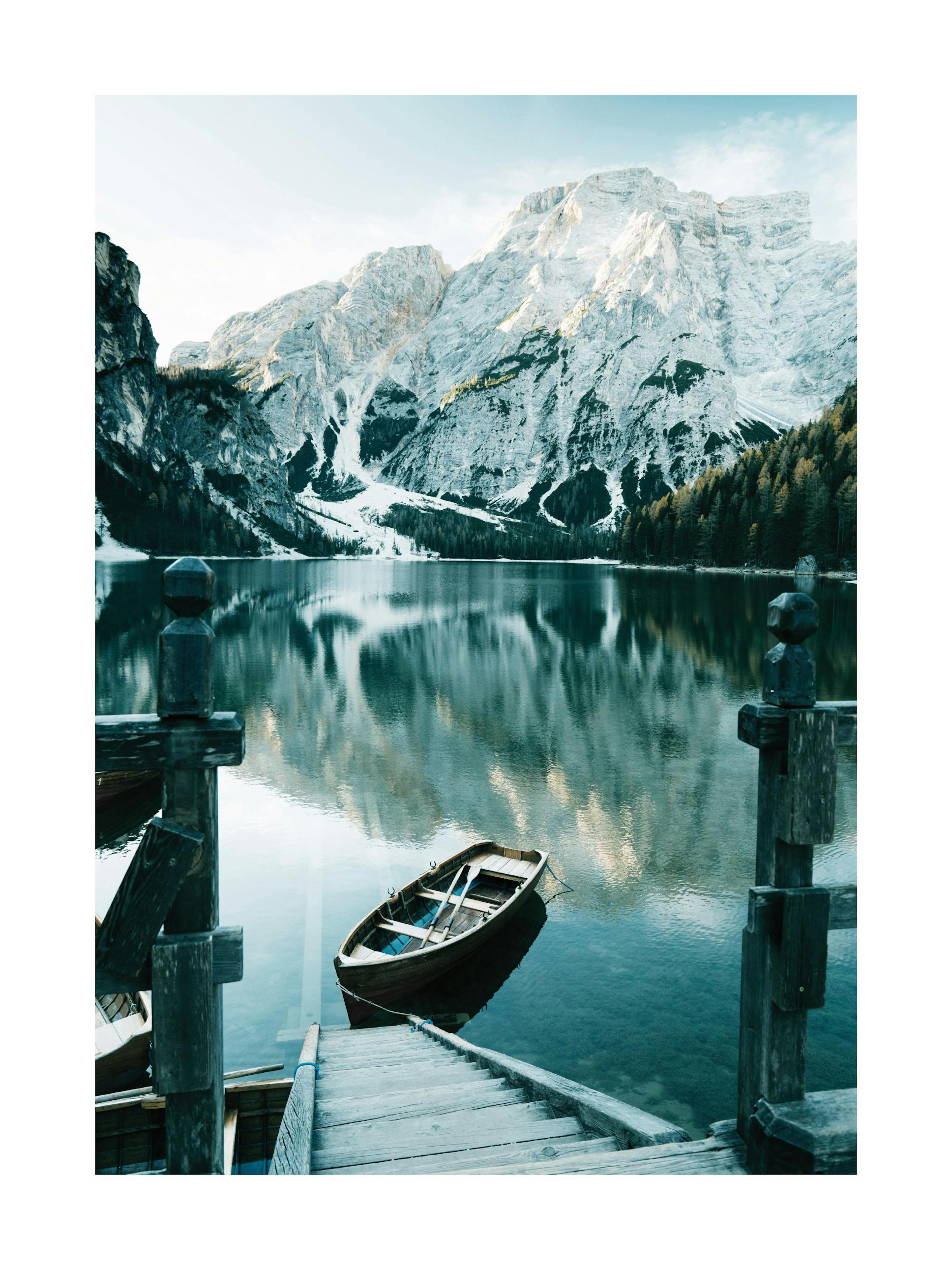 Photographic poster of an Alpine lake with a boat and wooden stairs, surrounded by snowy mountains and trees.