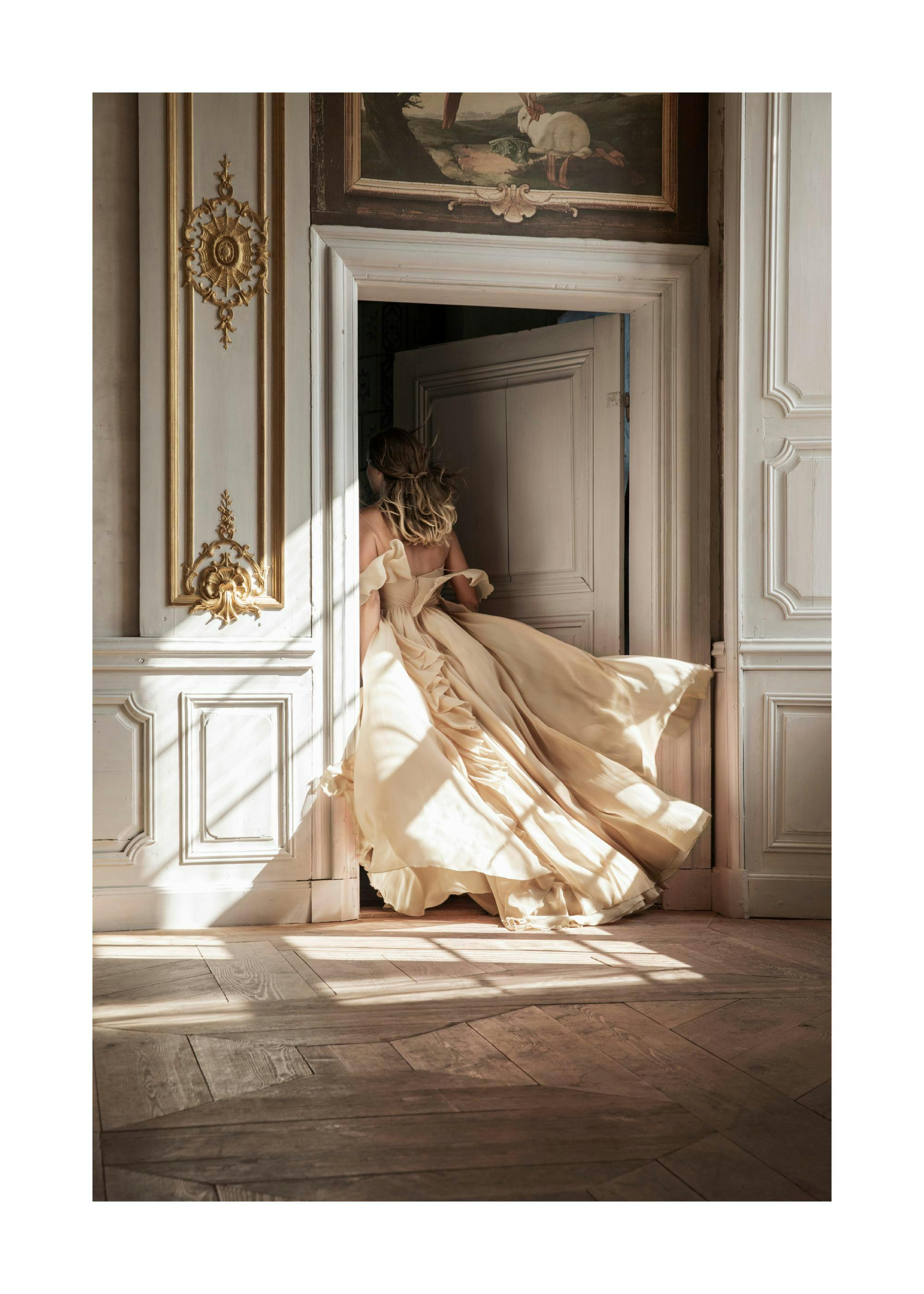 Fotografia de mulher em vestido bege, a entrar por porta barroca dourada, luz dramática e chão de madeira.