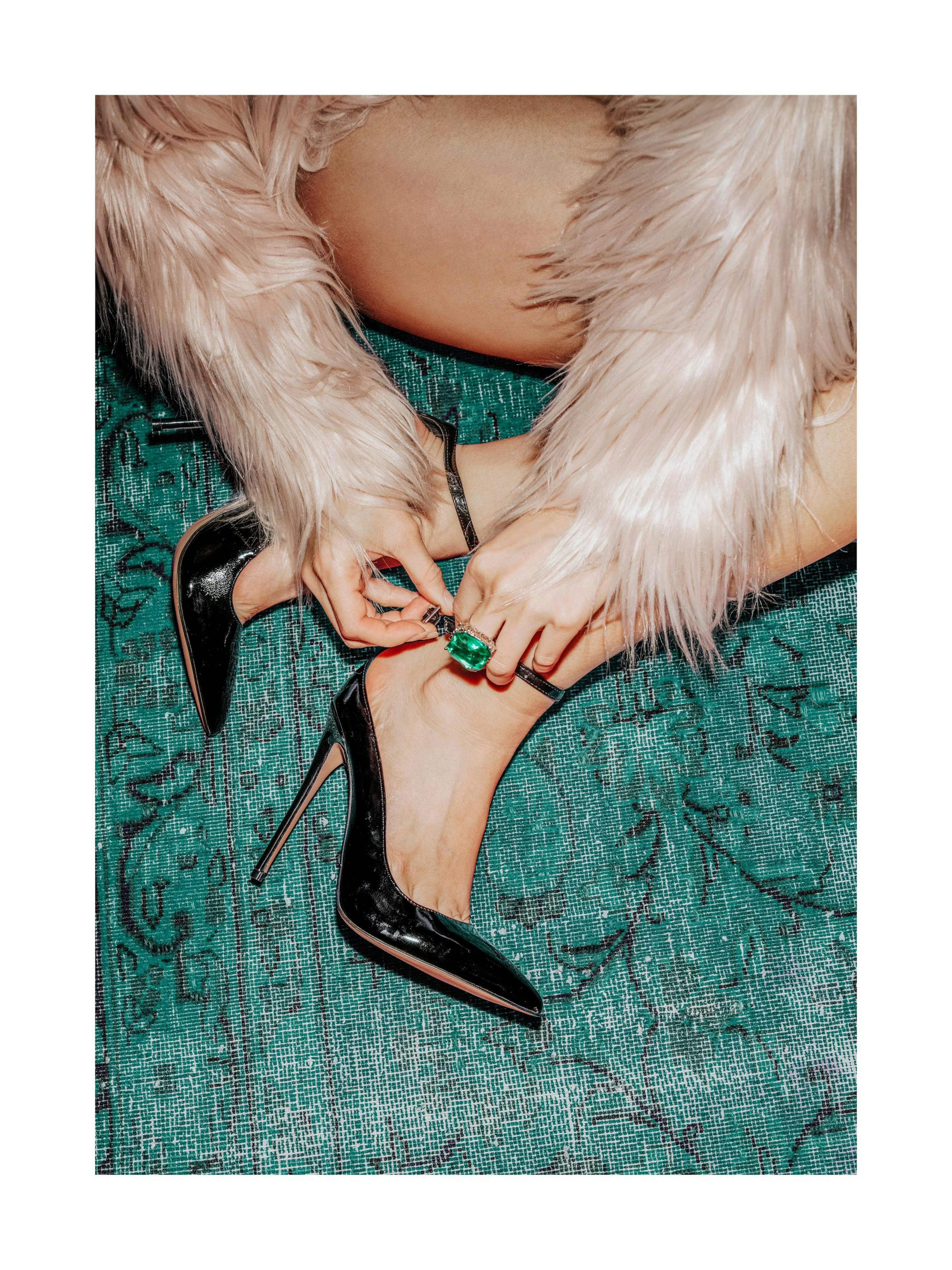 Póster de mujer abrochándose tacones negros con un anillo de esmeraldas y un abrigo de piel sintética rosa sobre una alfombra ve