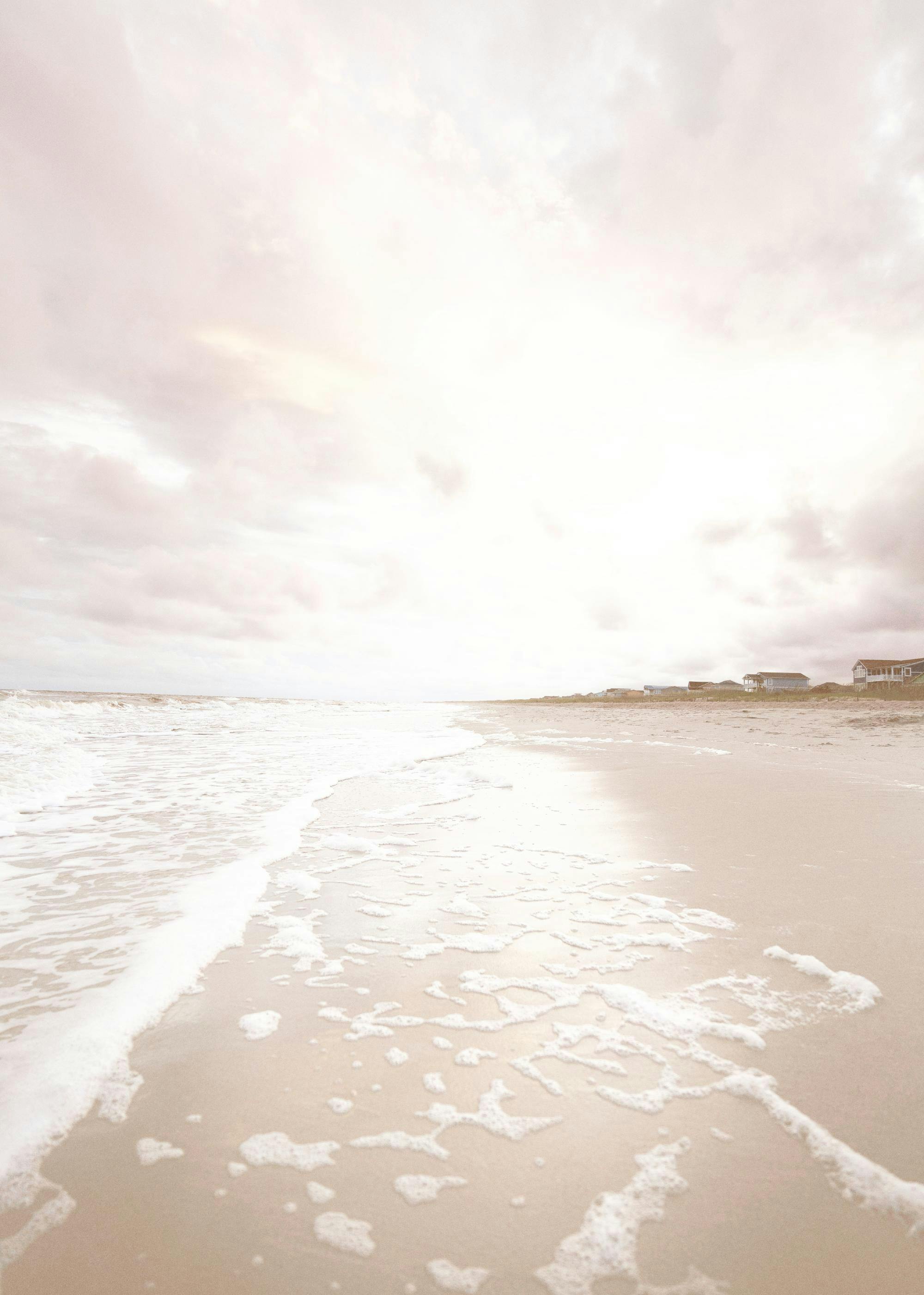 Póster de playa beige con espuma marina, casas costeras y cielo nublado, ideal para decoración de pared.