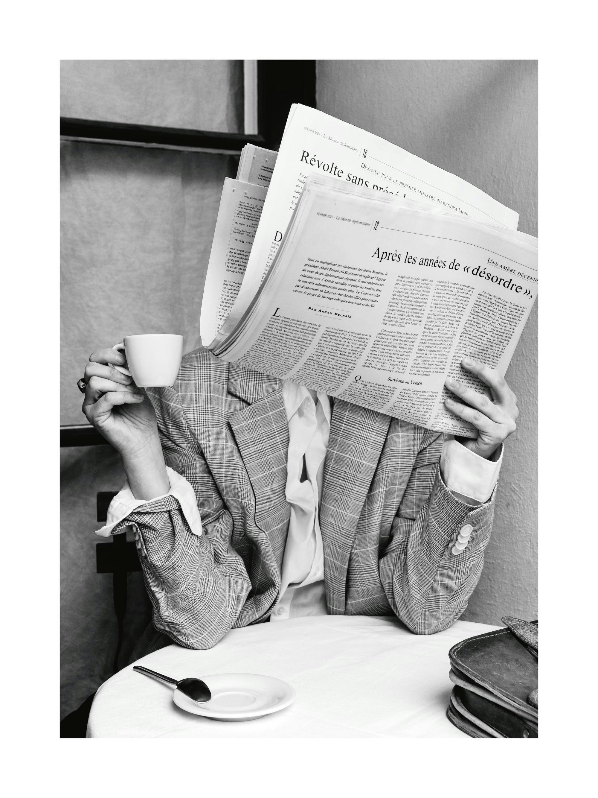 Svartvitt fotografi av en person som dricker kaffe och läser tidningen vid ett kafébord. Kaffeposter.