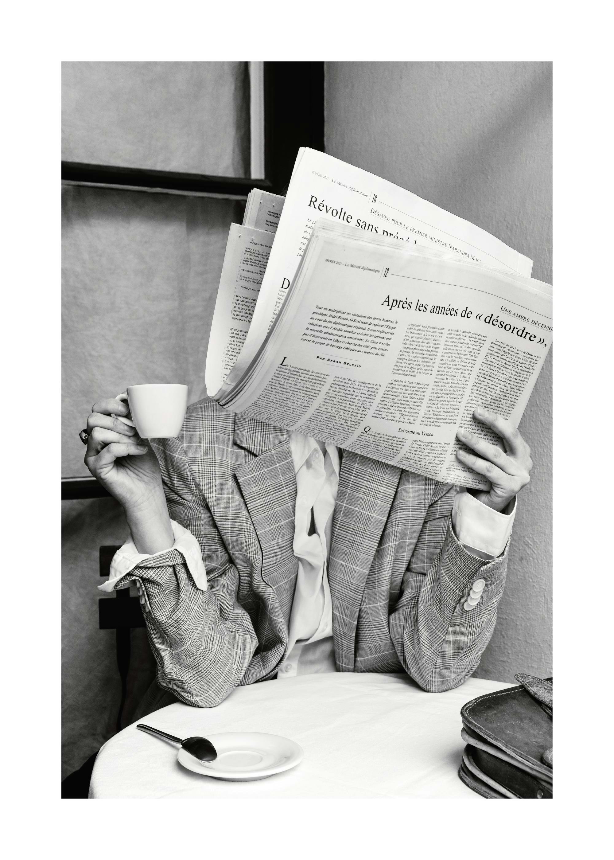 Black and white photography print of person with newspaper covering face, holding coffee cup at cafe table, poster