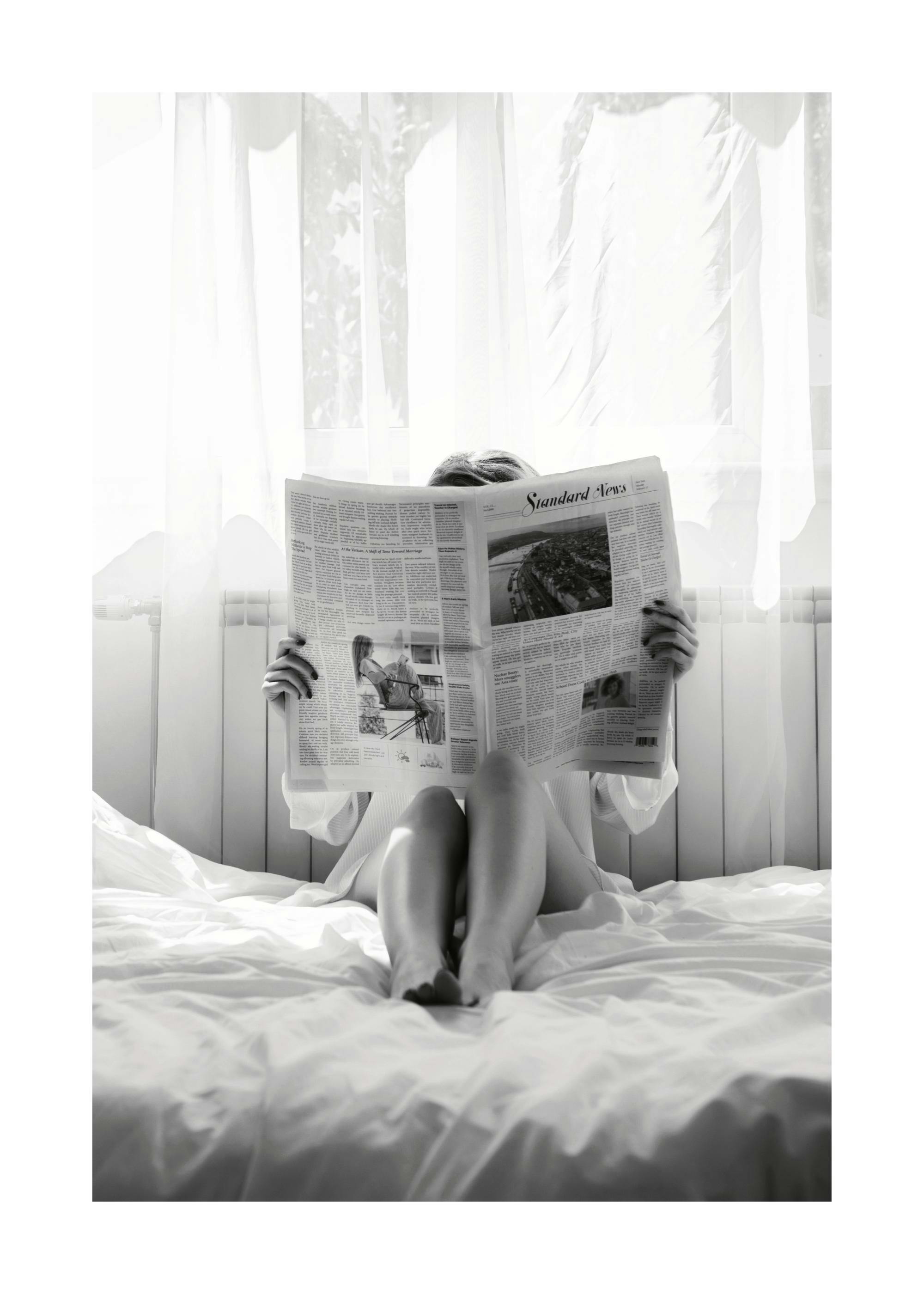 Svartvitt fotografi av en kvinna som läser tidningen i sängen, en klassisk och tidlös bild.