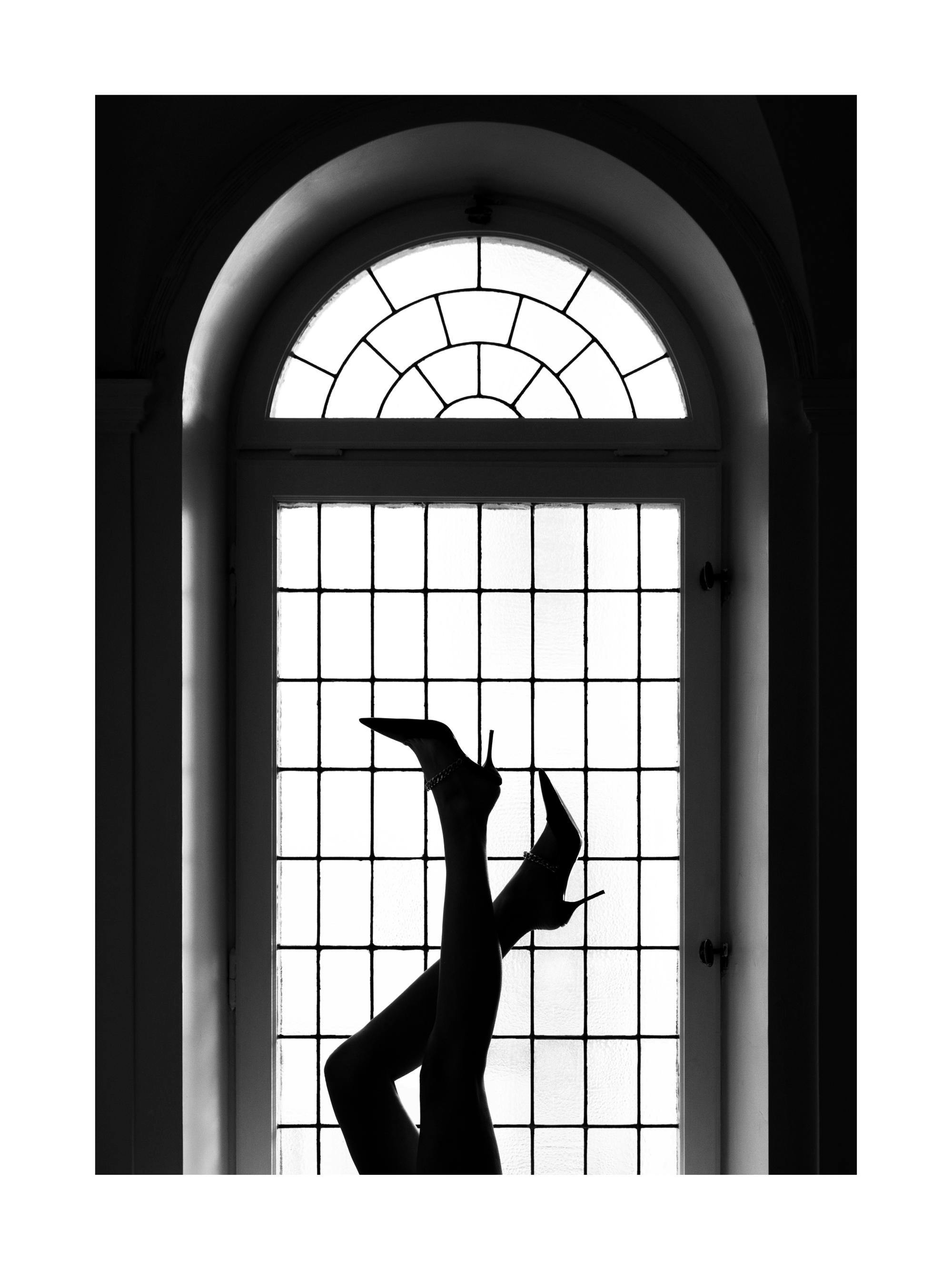 Svartvitt fotografi av ben med klackar mot ett välvt fönster, elegant och mystiskt.
