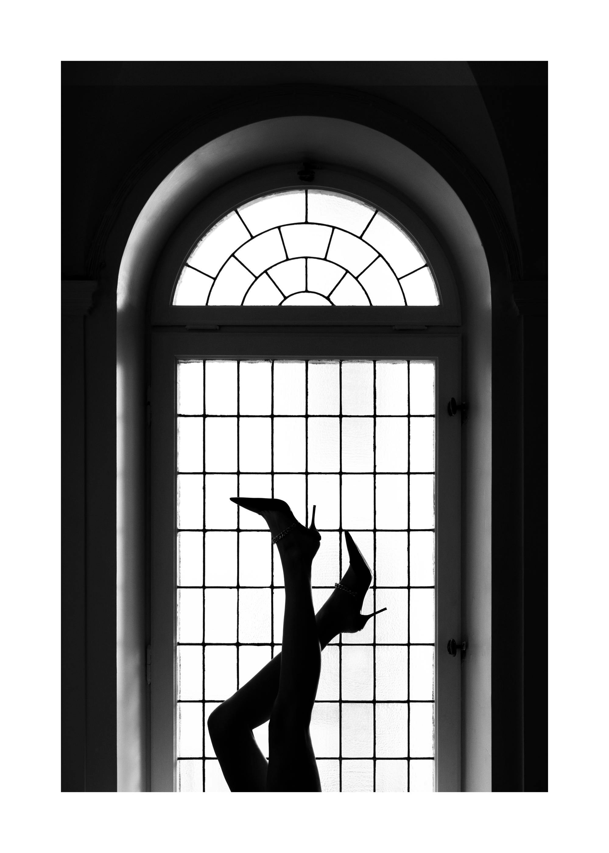 Black and white photograph of legs in high heels stretched upwards against an arched window, creating a striking silhouette.