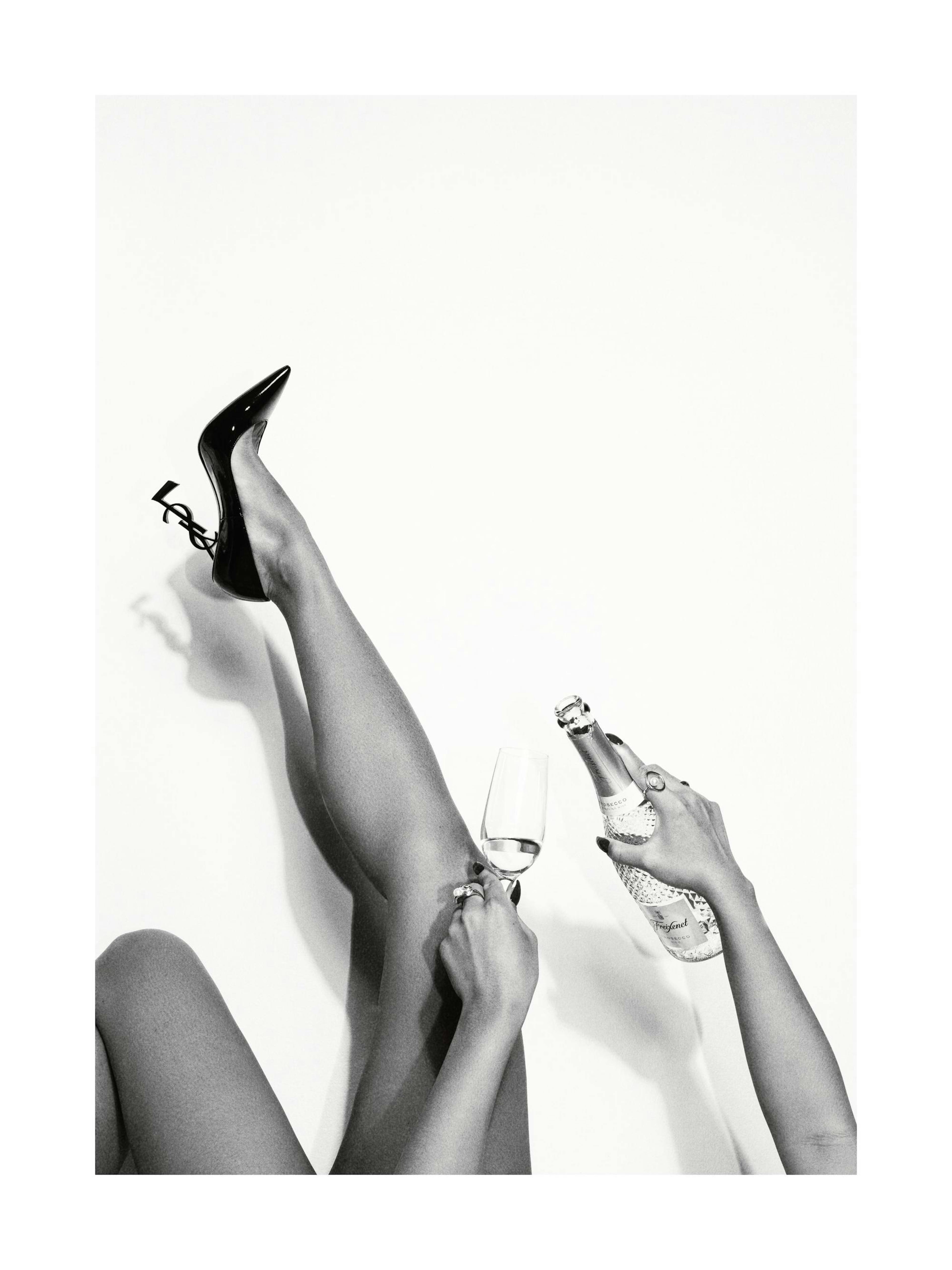 Schwarz-weißes Mode-Poster mit Beinen, die einen schwarzen Absatzschuh tragen, während Hände Champagner in ein Glas füllen.
