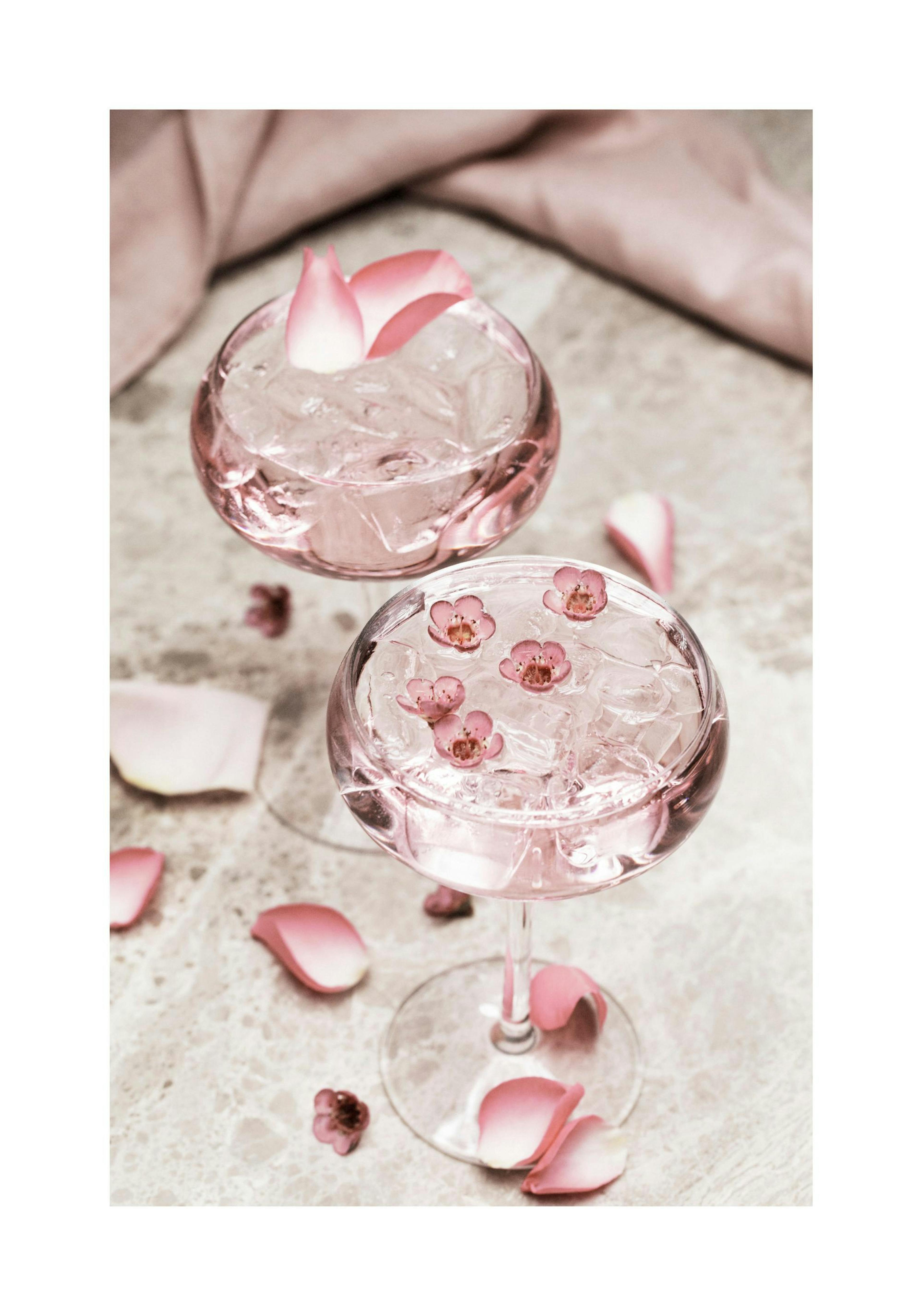 Fotografie von zwei rosa Cocktails mit Eis, verziert mit Rosenblättern und Blüten, auf Marmoruntergrund.