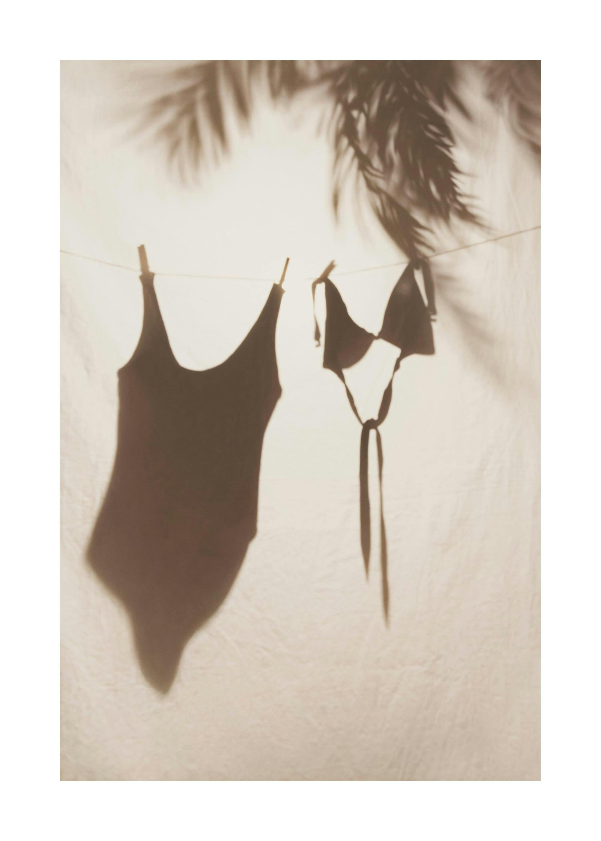 Shadow of black swimwear and bikini top drying on a line, with palm tree shadows above, on a light background.