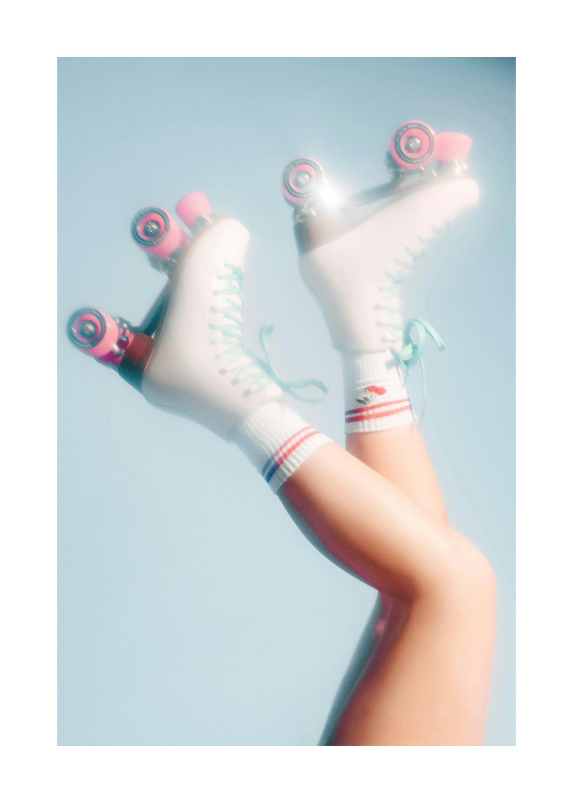 Fotografía de dos piernas con patines de ruedas blancos y rosas, con el cielo azul de fondo y un reflejo de sol.