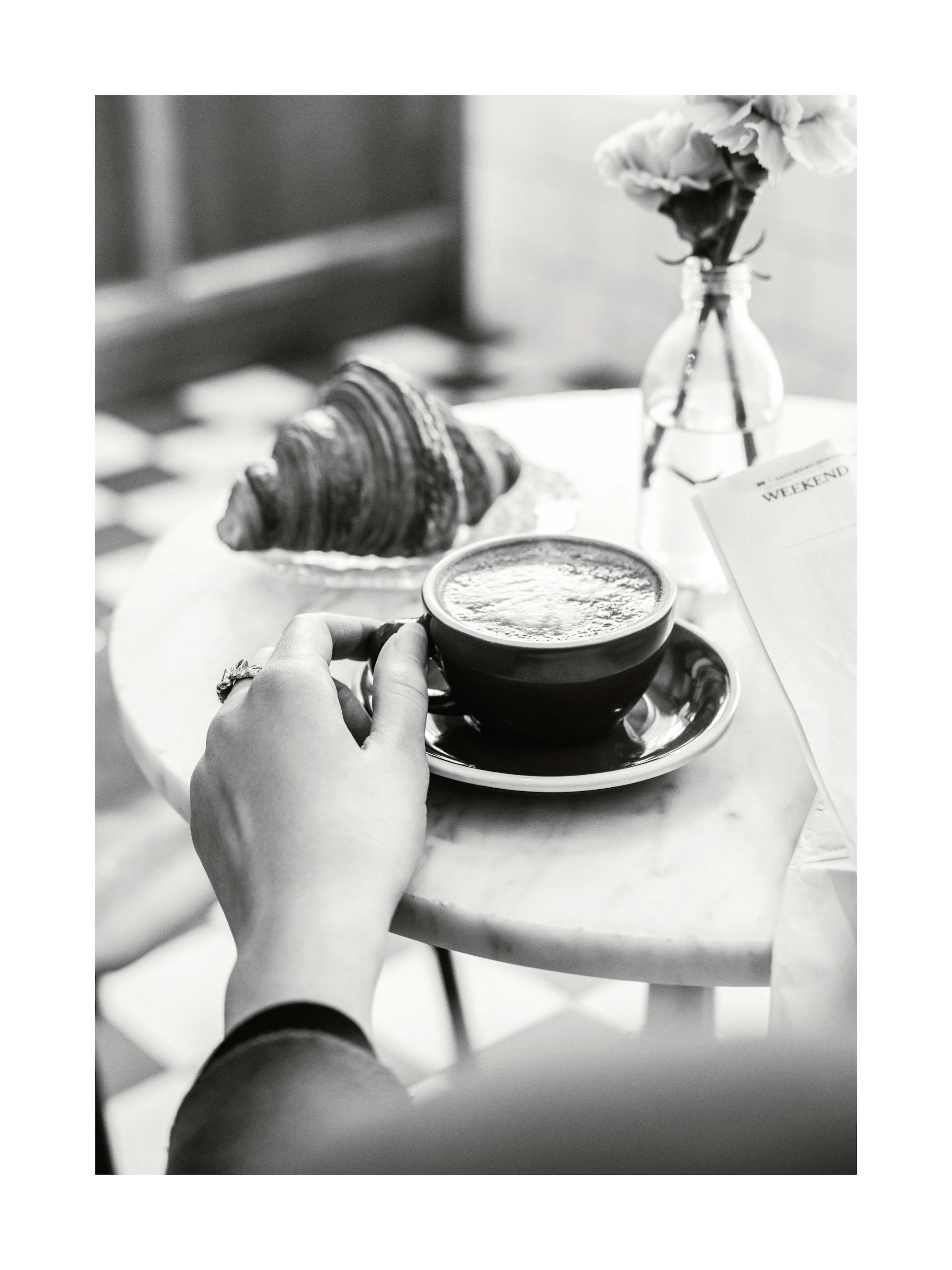 Affiche noir et blanc dune main tenant une tasse de café sur une table de petit-déjeuner avec croissant et fleurs