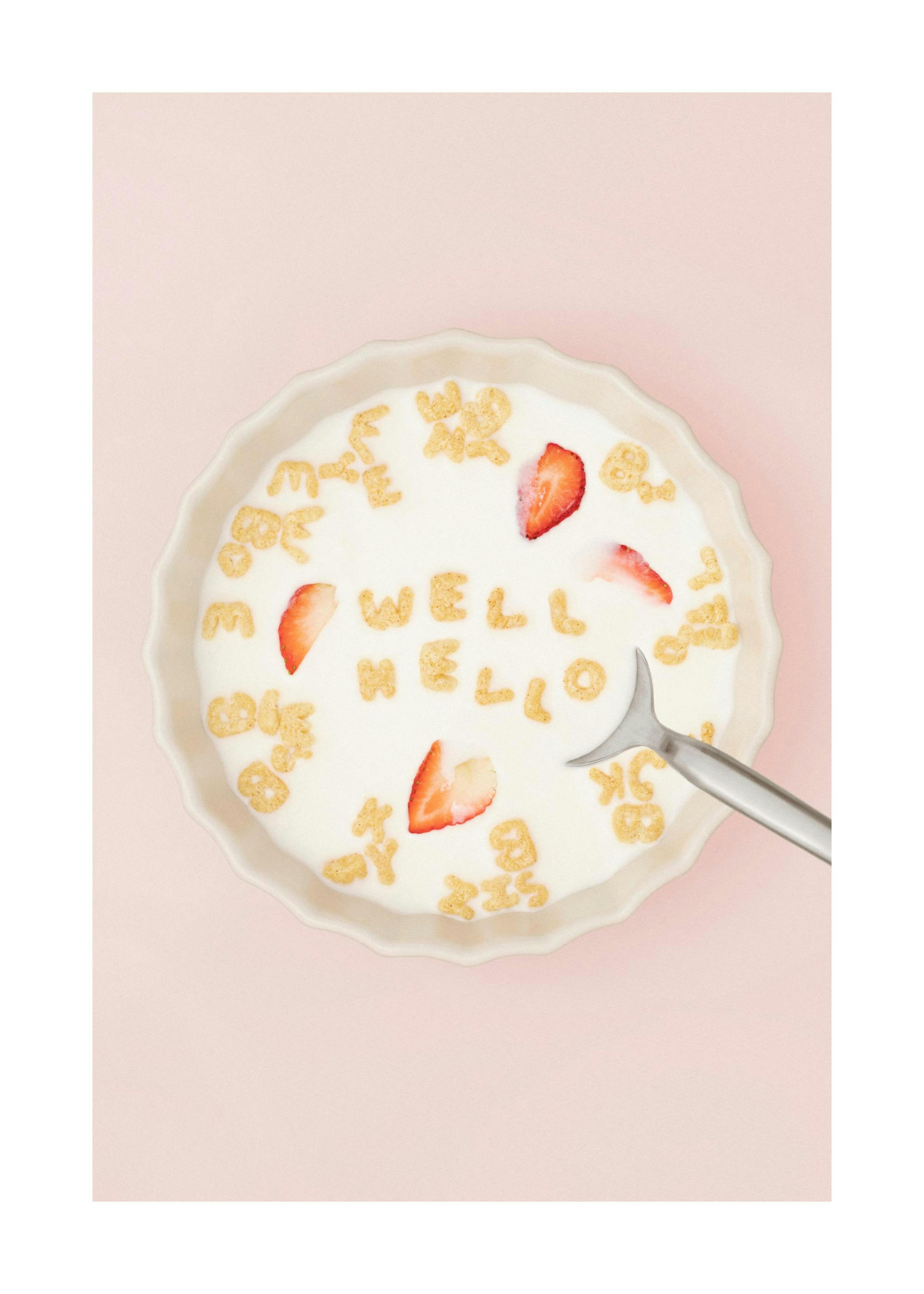 Tazón de leche con cereales de letras que forman Well Hello y fresas en un fondo rosa, ideal para decoración de cocina.