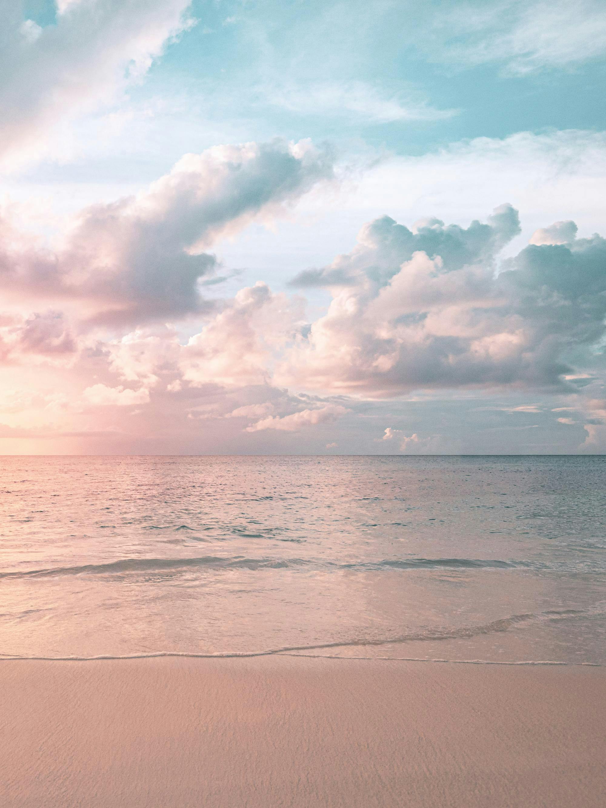 Ranta-auringonlasku pilvisellä taivaalla, jossa siniset ja vaaleanpunaiset sävyt kohtaavat. Aaltojen äärellä hiekkaranta.