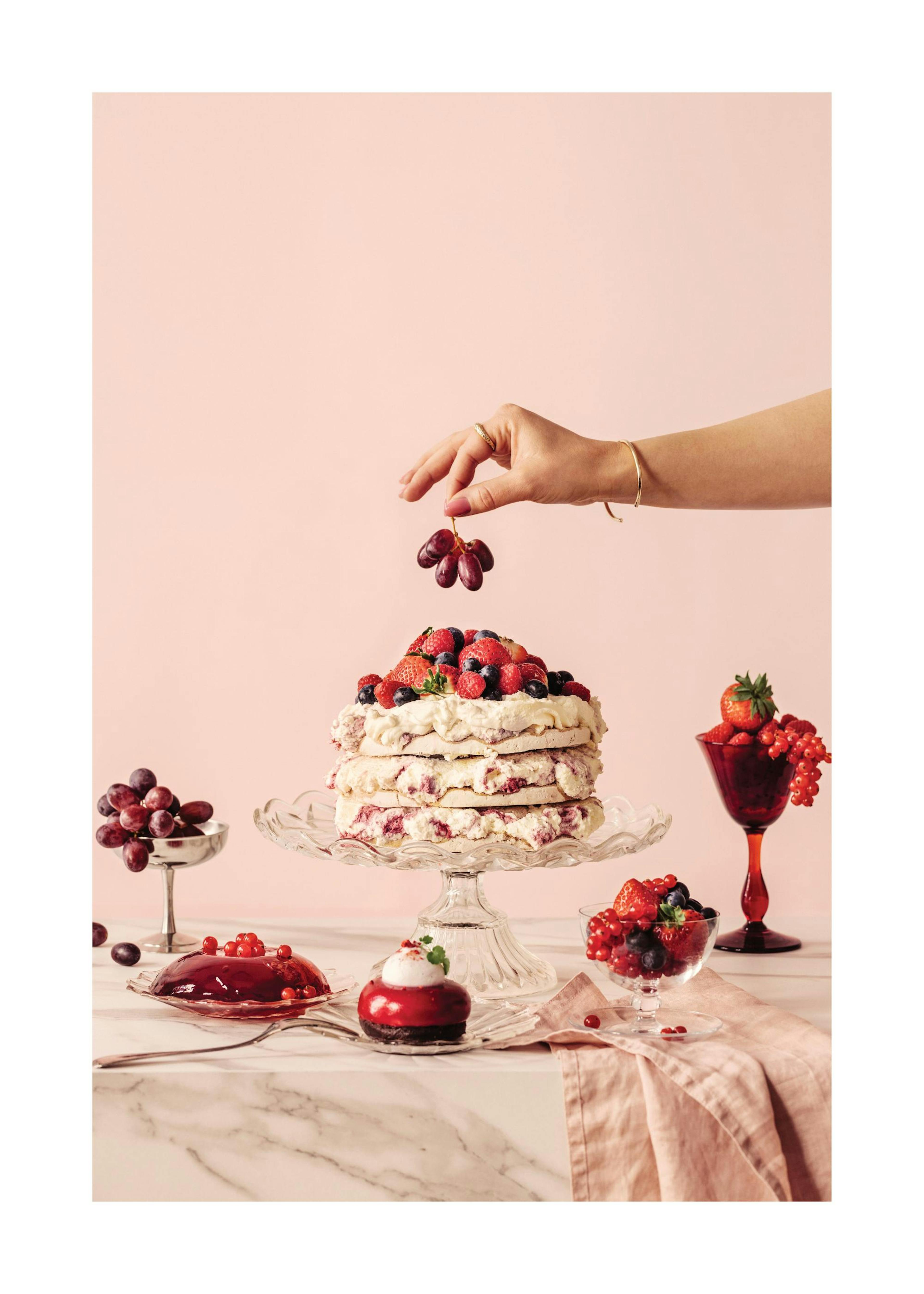 Hand adding grapes to a berry cake on a table with other desserts against a pink background.