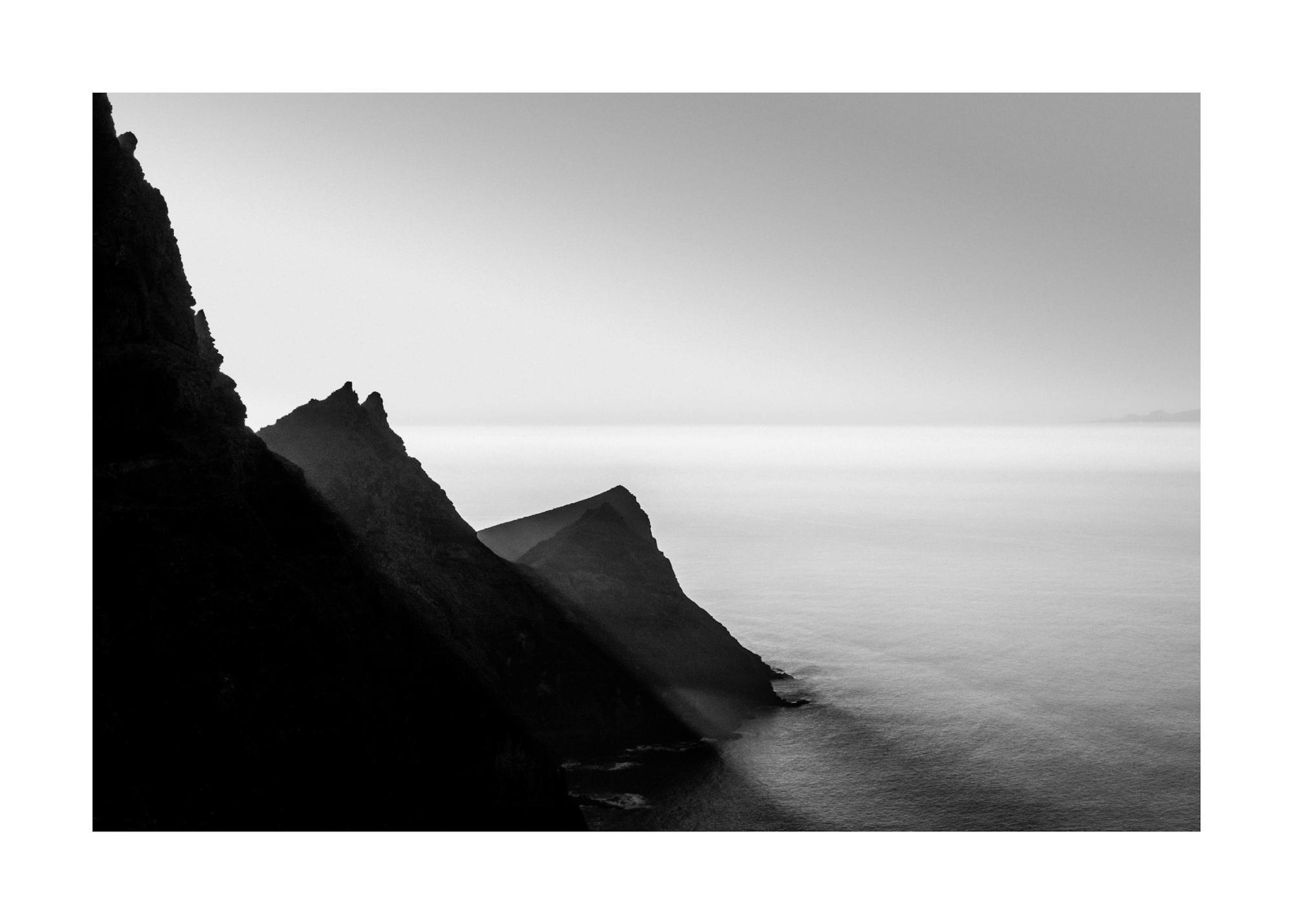 Black and white photography of dark, jagged mountains meeting a calm, hazy sea and horizon.