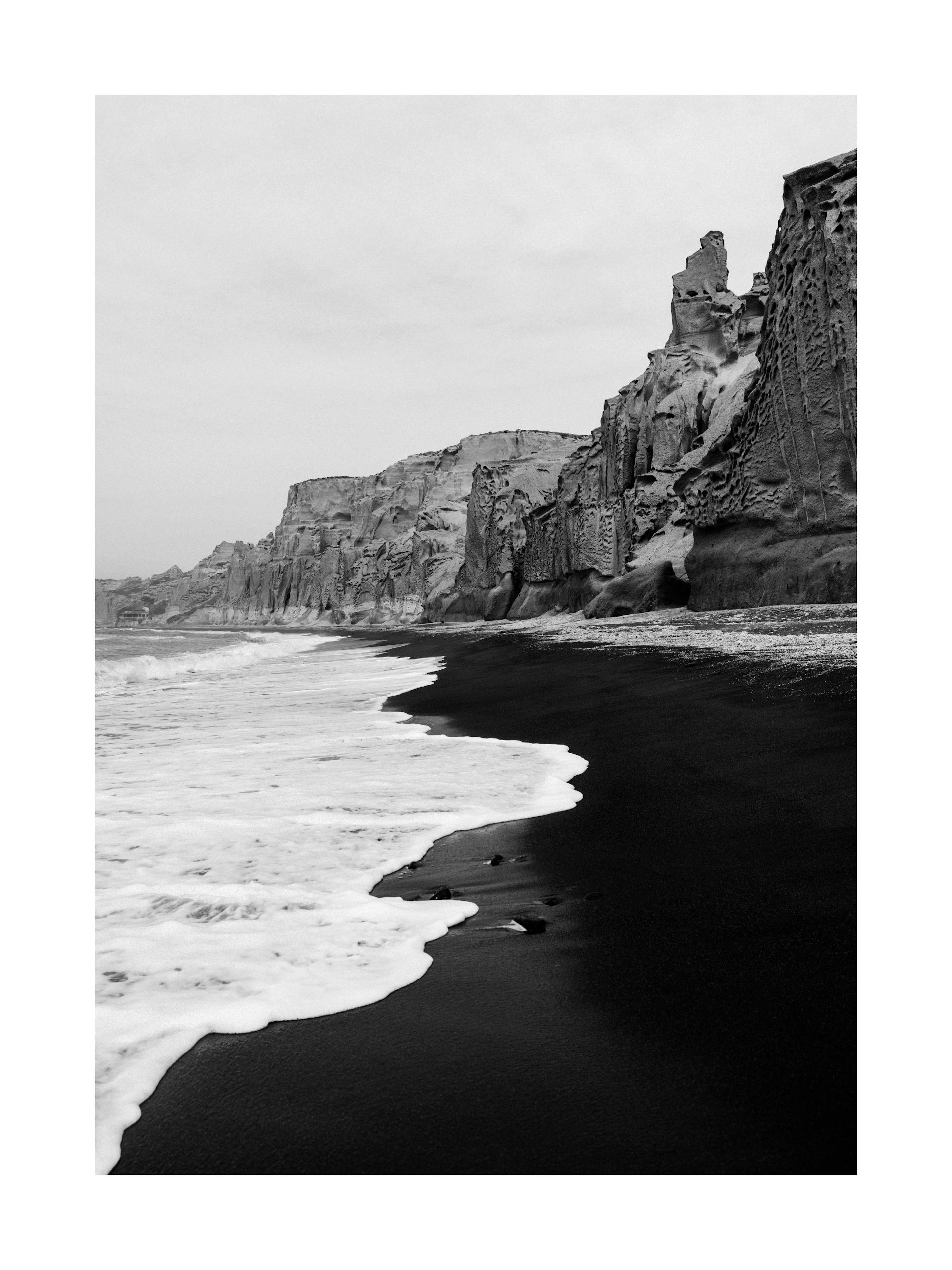 Sort-hvidt fotografi af en mørk strand med bølger og høje klipper under en overskyet himmel.