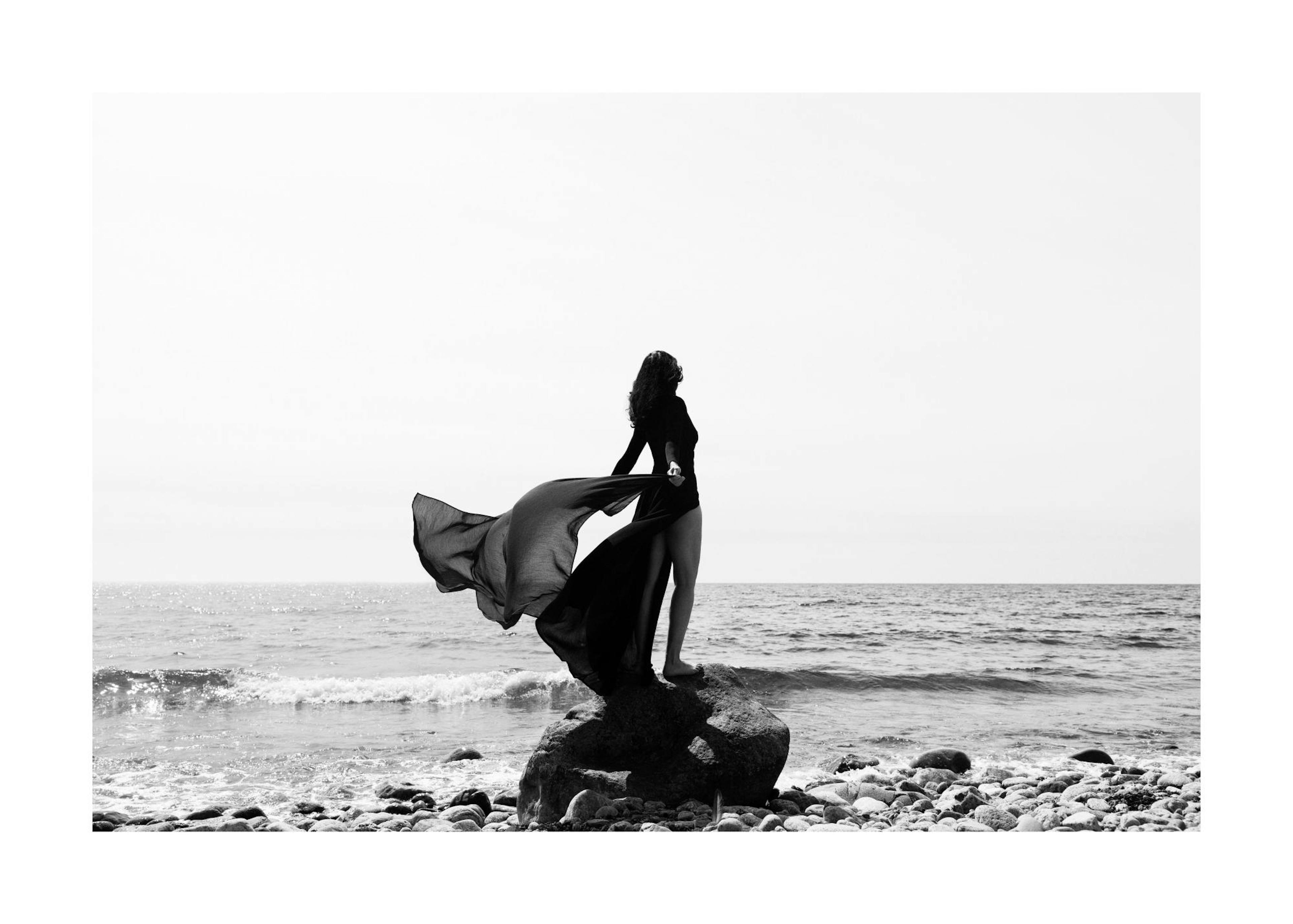 Black and white photo of a woman in a flowing dress standing on a rock in the water, dramatic and calming