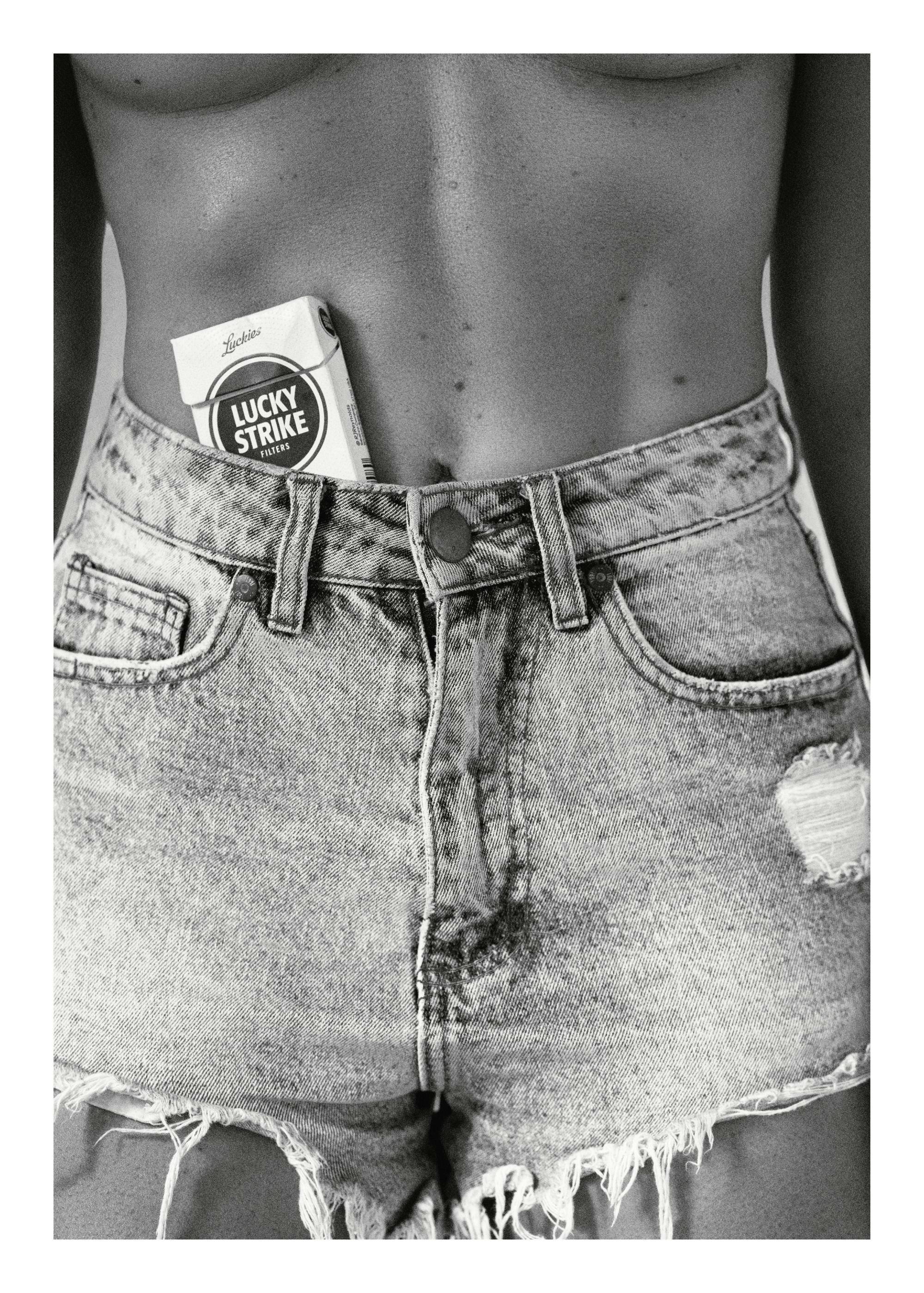 Zwart-wit poster van een vrouw in denim short met een pakje Lucky Strike sigaretten in haar tailleband.