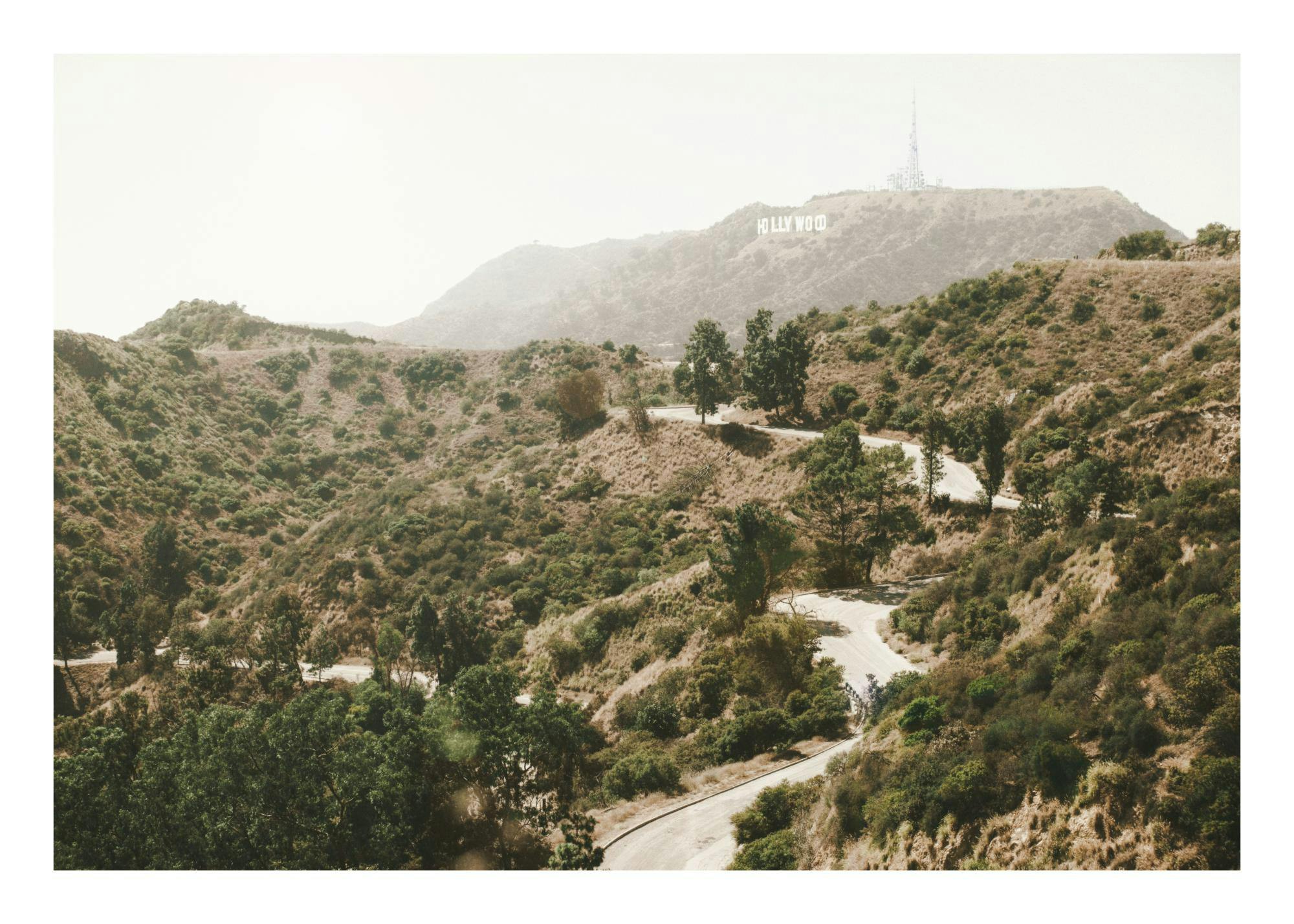 Poster strada tortuosa tra colline verdi, cartello Hollywood in lontananza, cielo azzurro limpido, paesaggio