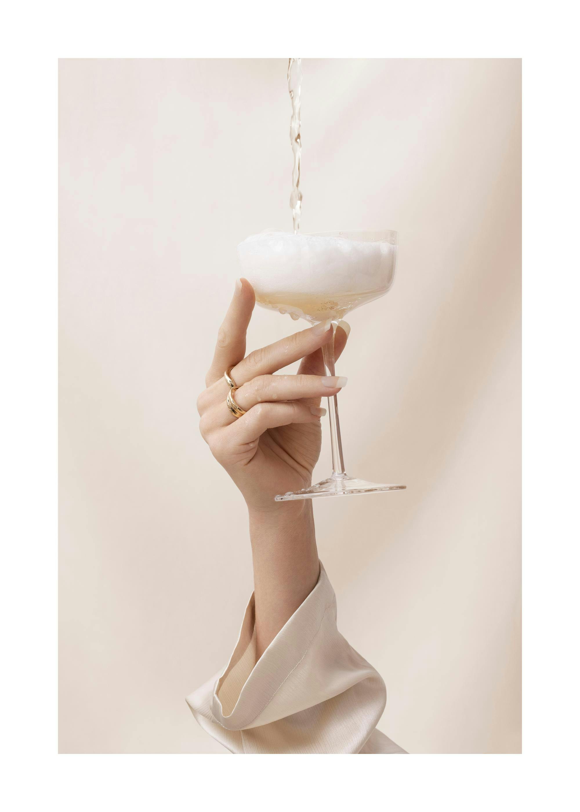Kvinnoarm som håller ett champagneglas med champagne som hälls i, mot en beige bakgrund. Festlig konst.