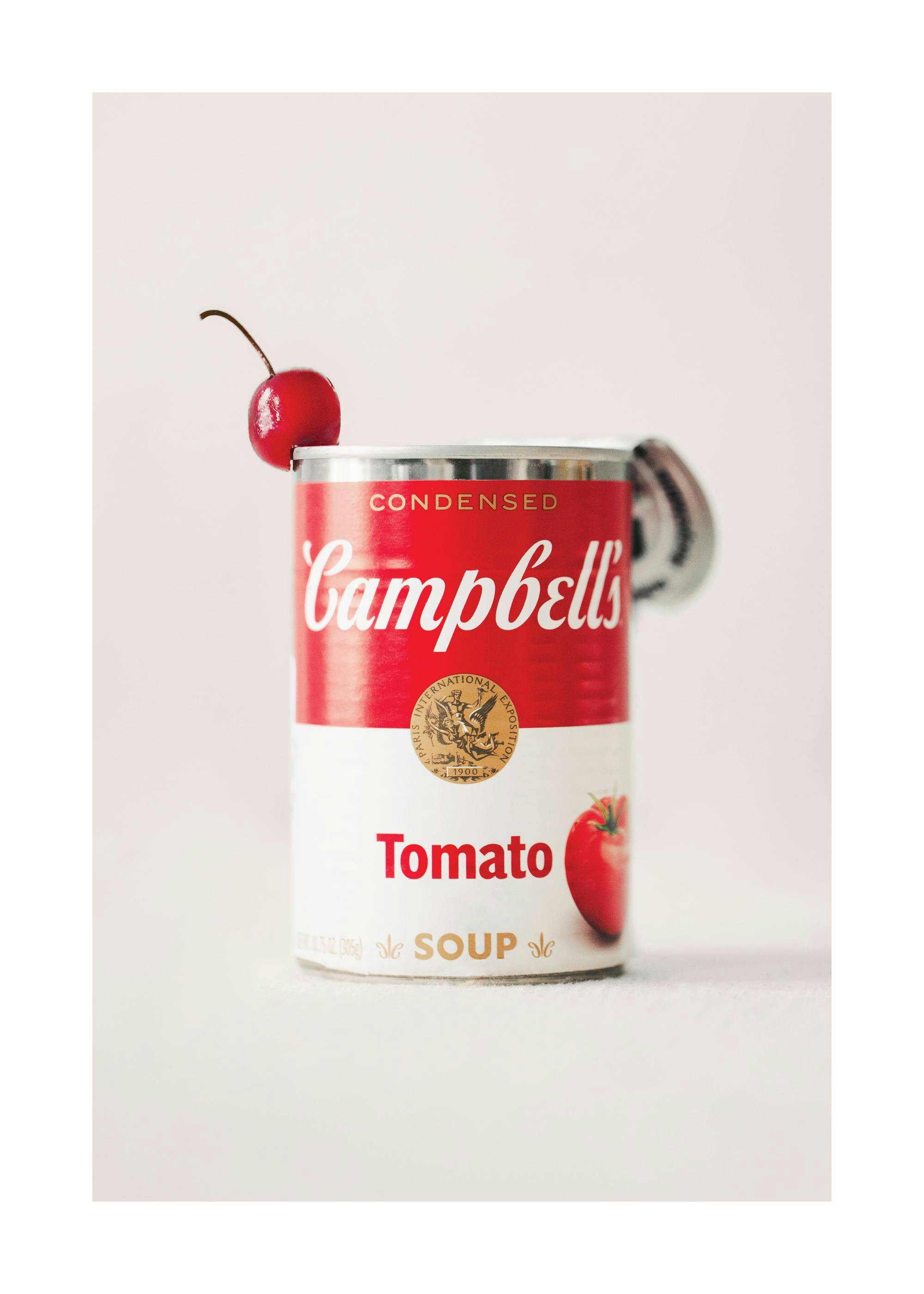 Retro photograph of a red and white tomato soup can with a cherry on top, against a beige background.