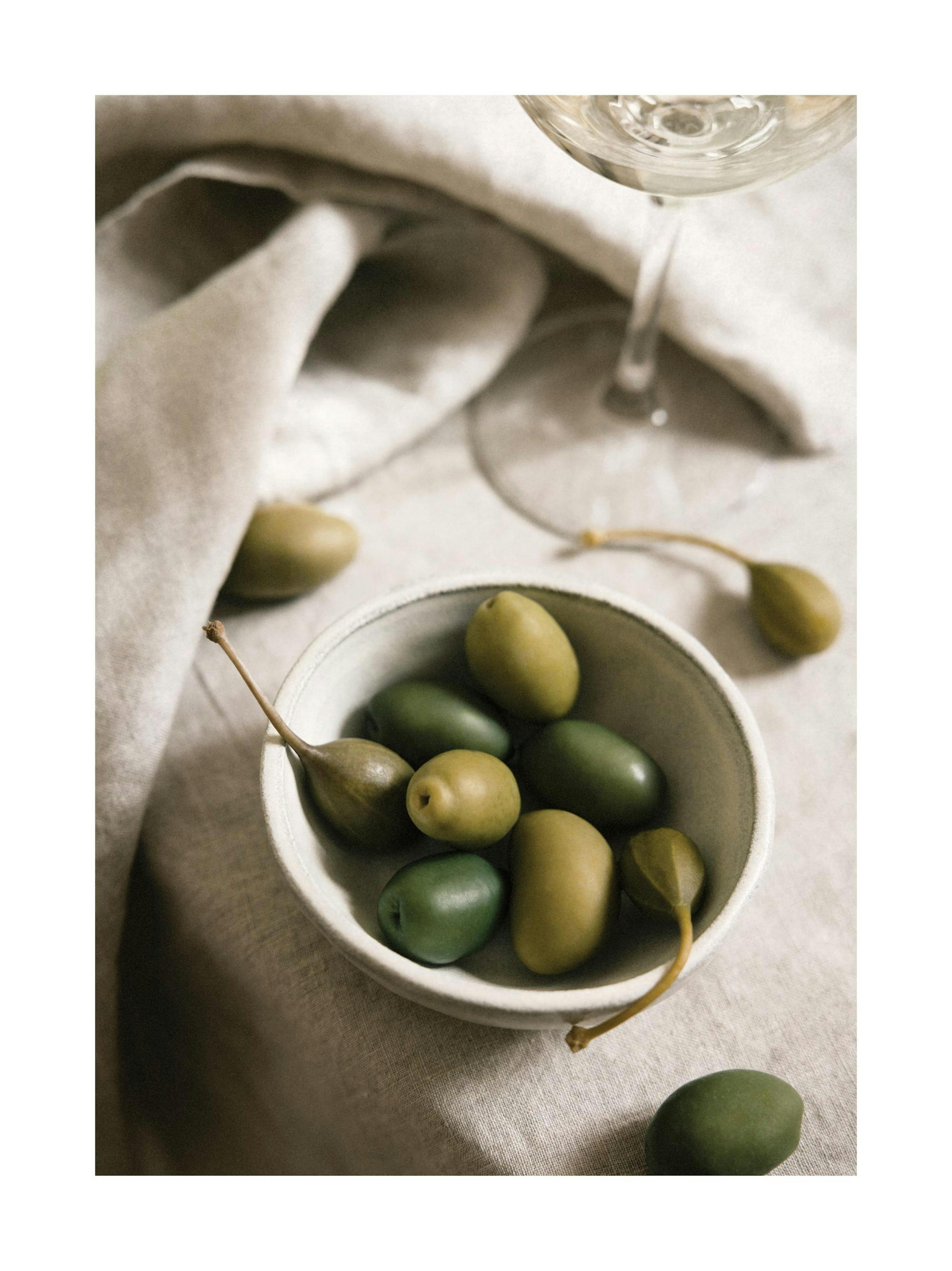 Bowl of green olives and capers on a linen tablecloth, with a glass of white wine in the background.