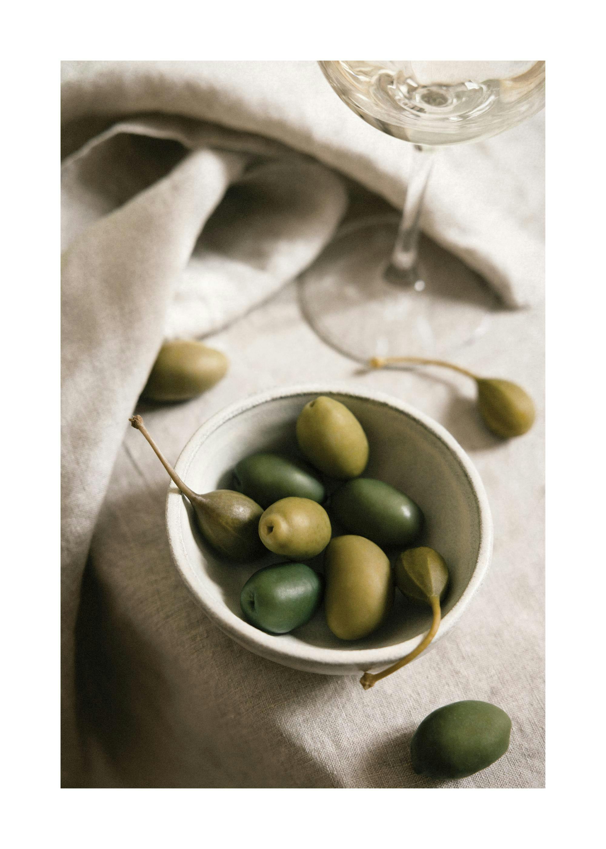 Affiche dun bol dolives vertes et de câpres, avec un verre de vin blanc sur une table en toile texturée.