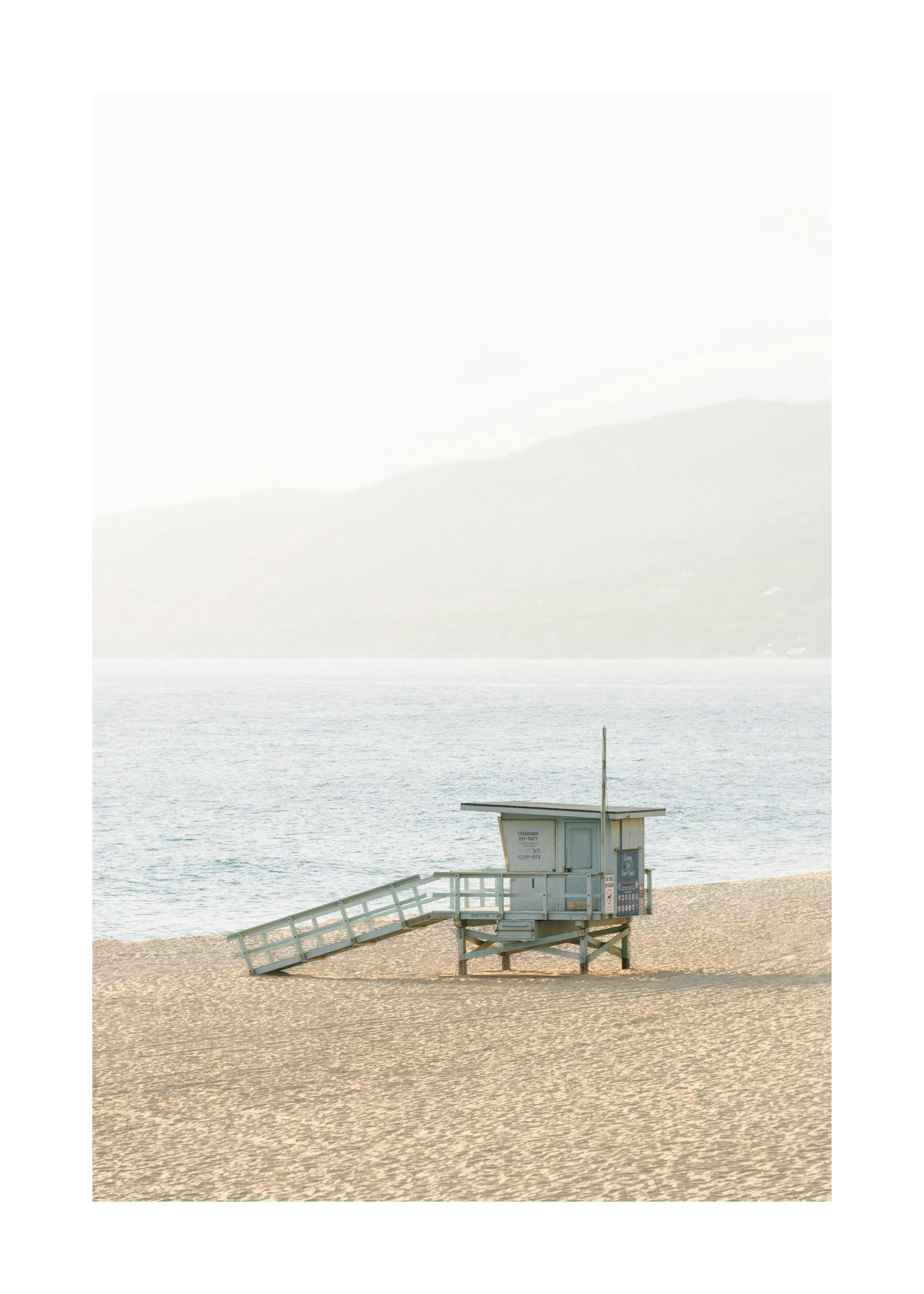 Malibu beach photo print of a turquoise lifeguard tower on sand with the ocean and mountains in the background.