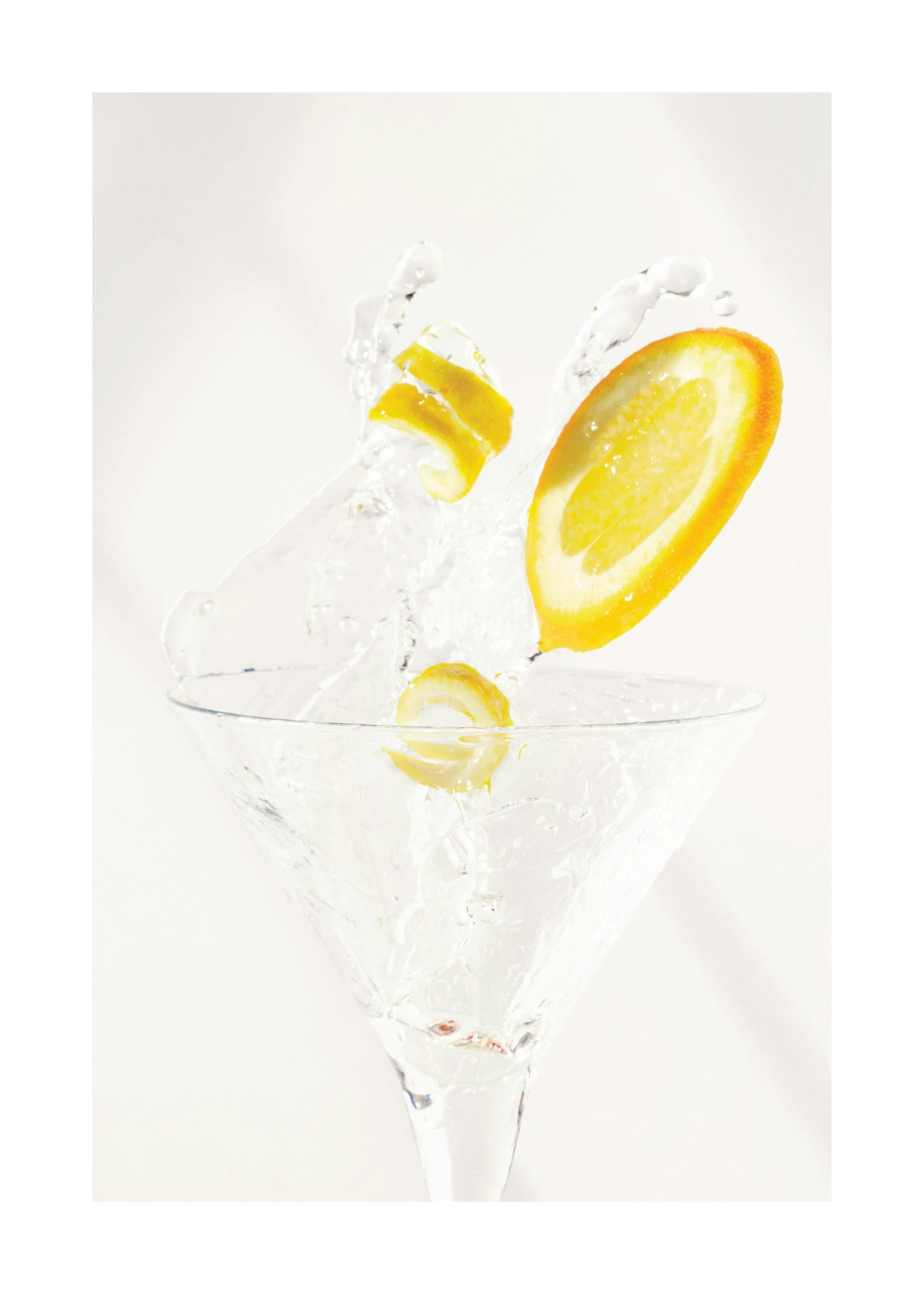 Orange slice and lemon peel splashing into a cocktail glass, clear liquid, white background.