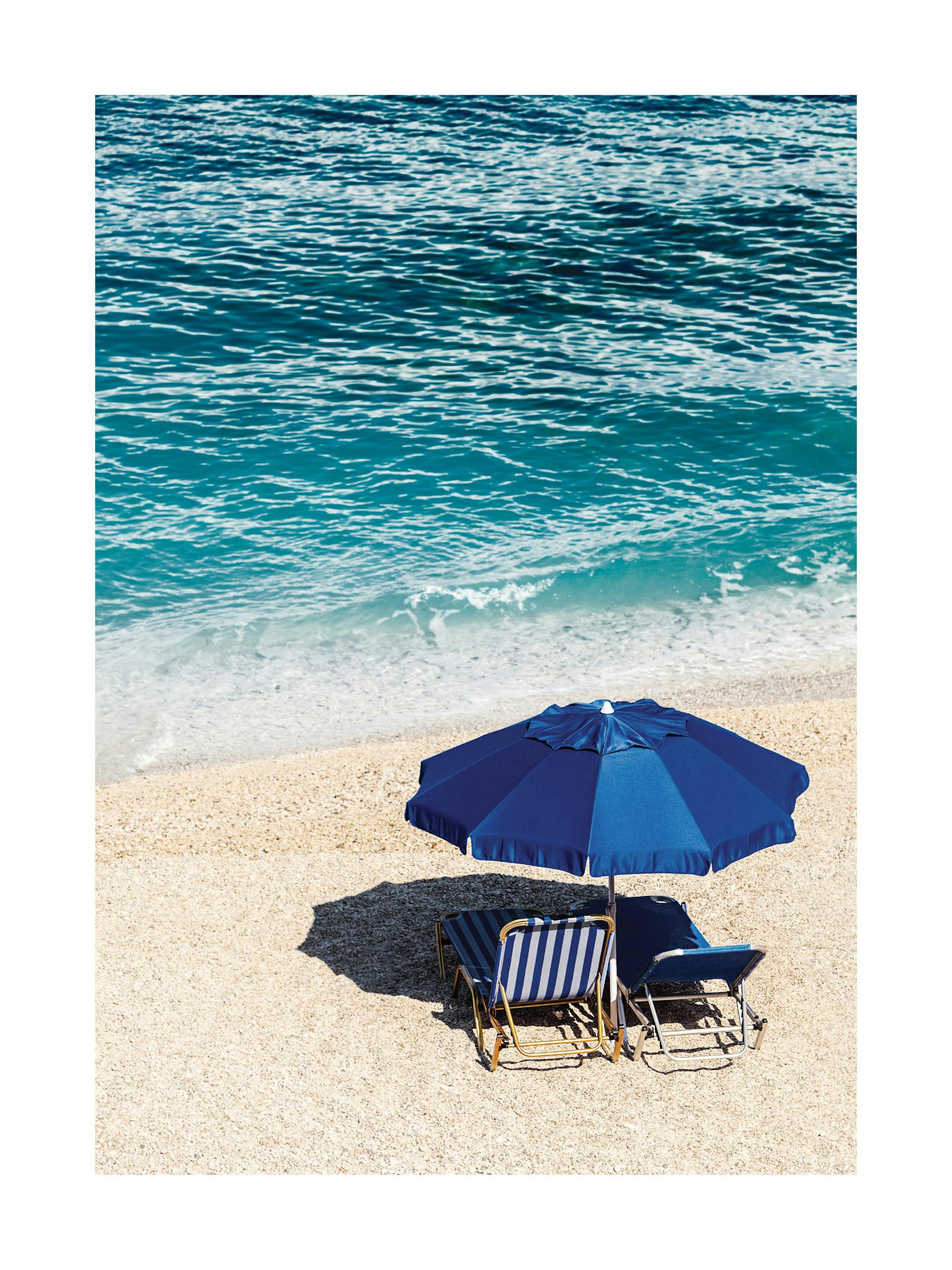 Blue parasol and striped sunbeds on a white sand beach with clear blue ocean in a poster print.