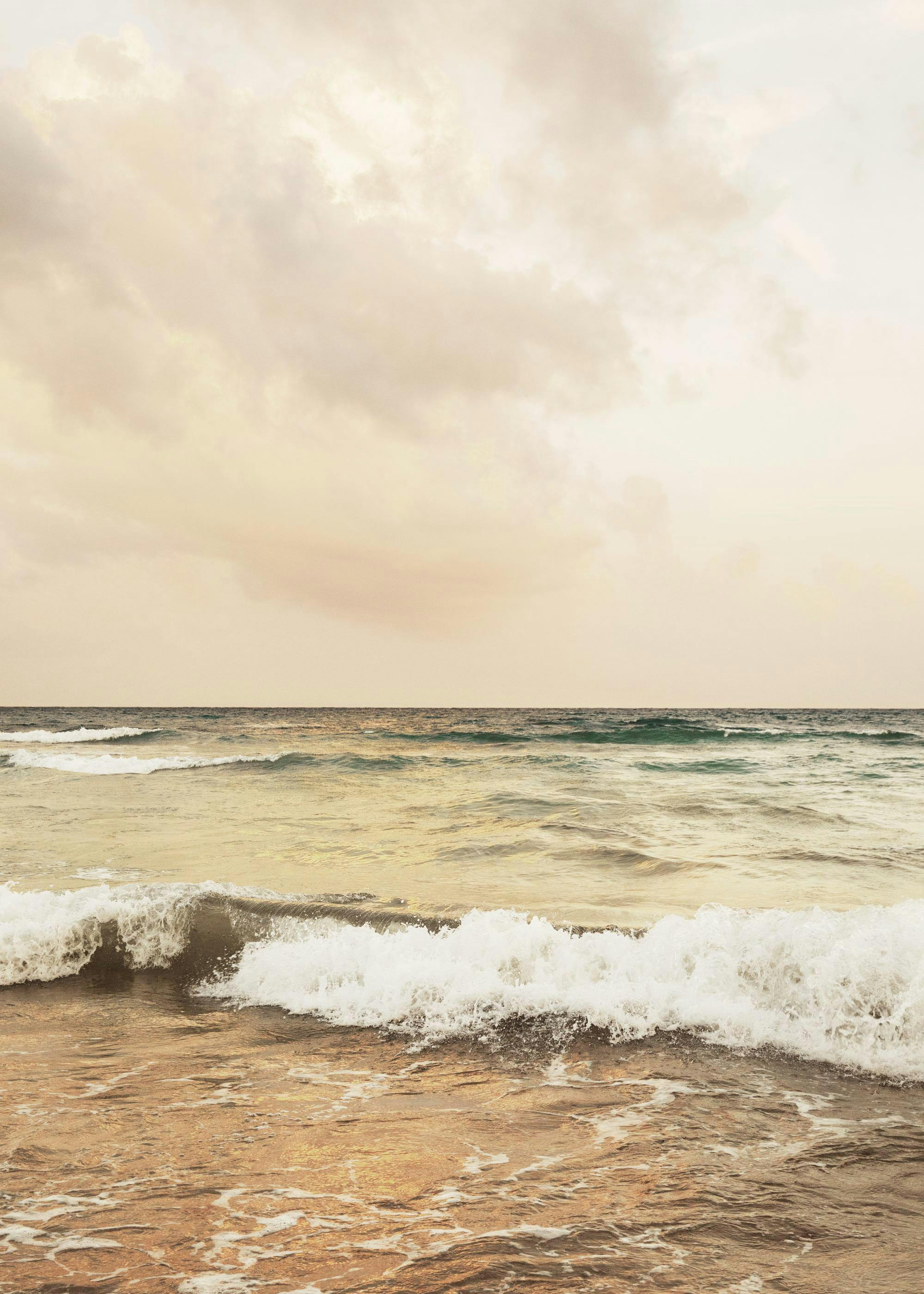 Ocean at sunset with soft clouds and gentle waves breaking on the shore, captured in a warm, golden photographic print.