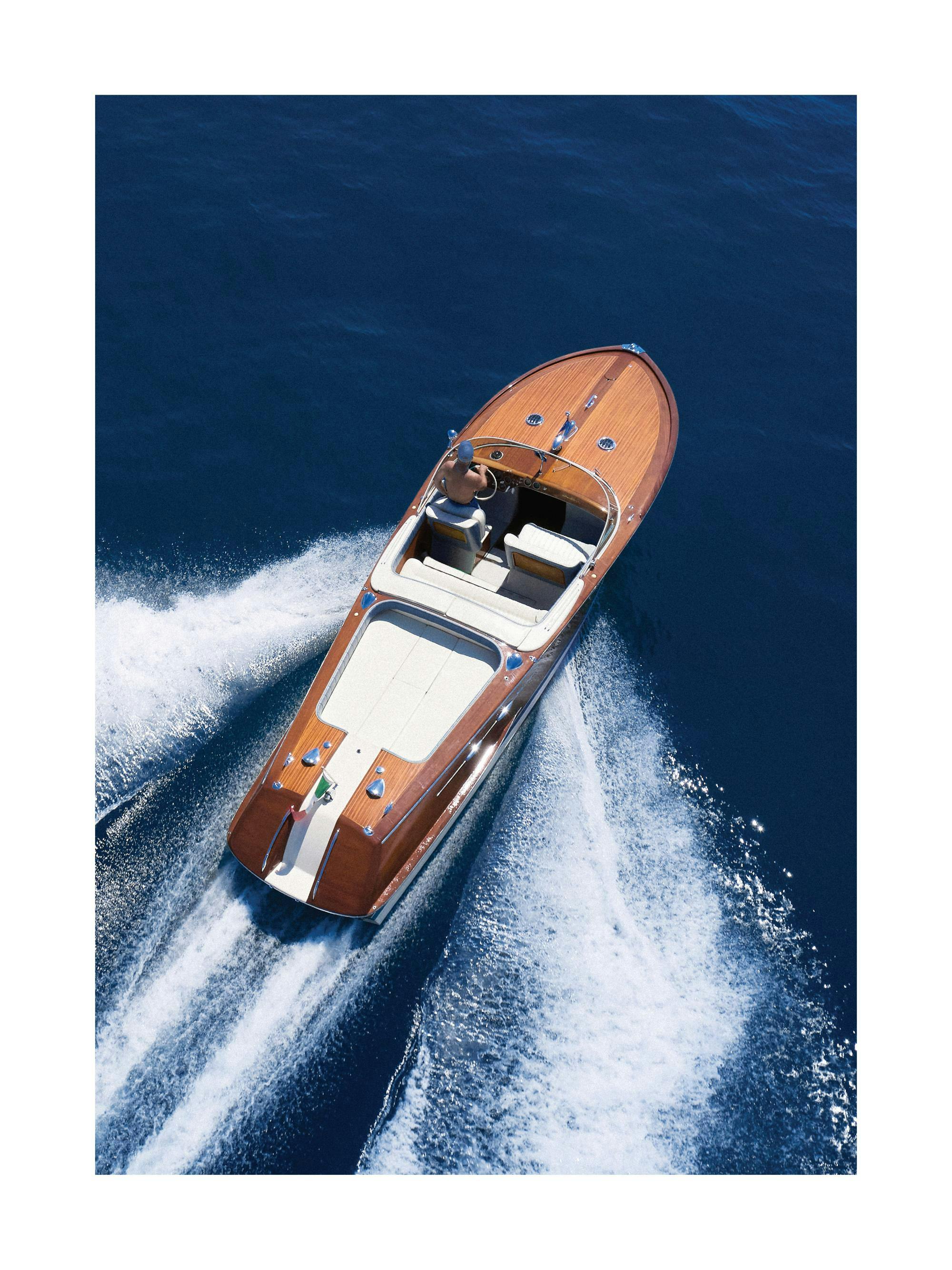 Luftfoto av en klassisk motorbåt i trä som skär genom mörkblått vatten, vilket skapar vita vågor bakom sig.