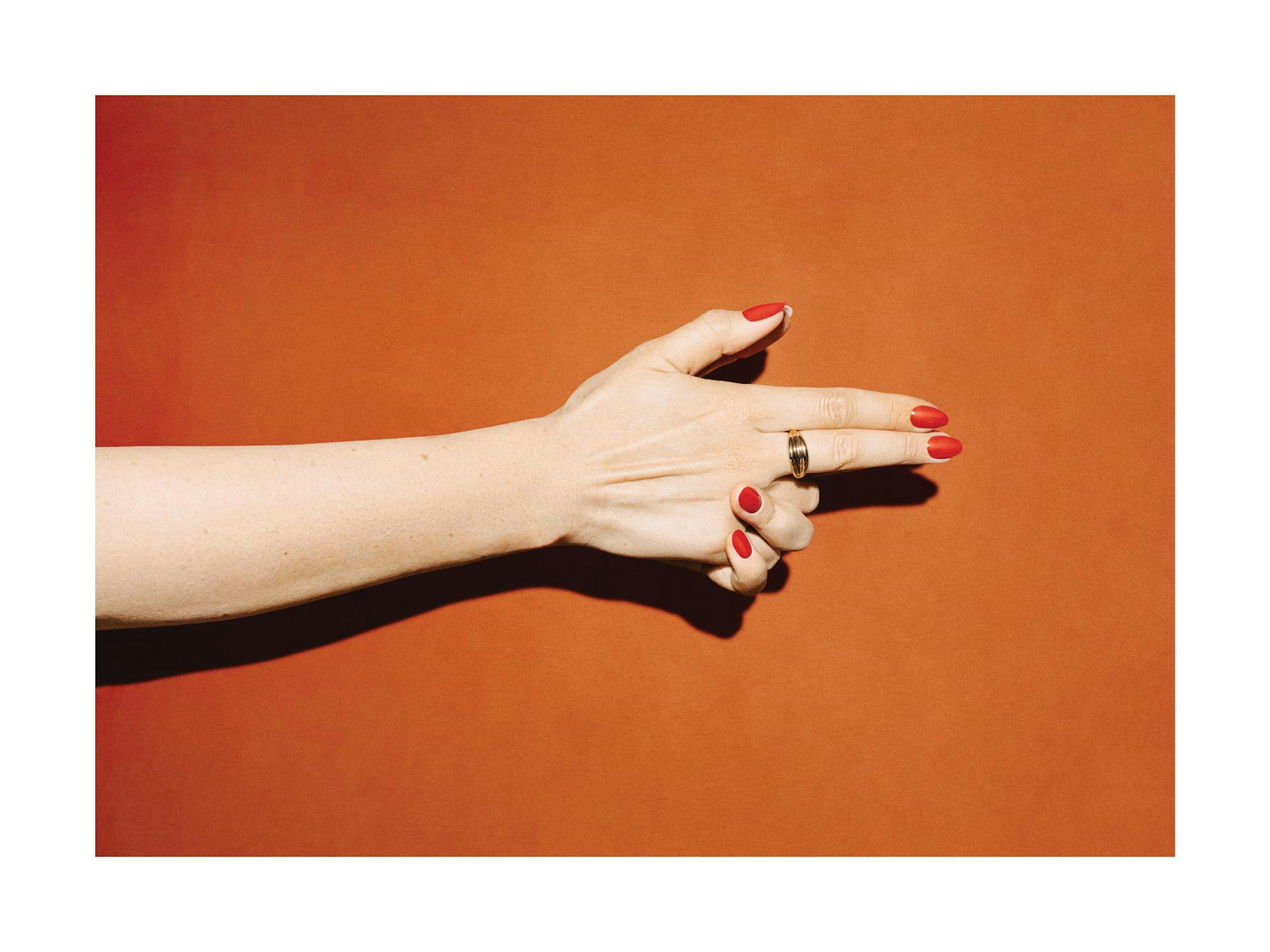 Mano femminile con unghie rosse che forma una pistola con le dita su sfondo arancione vibrante, con un anello dorato.