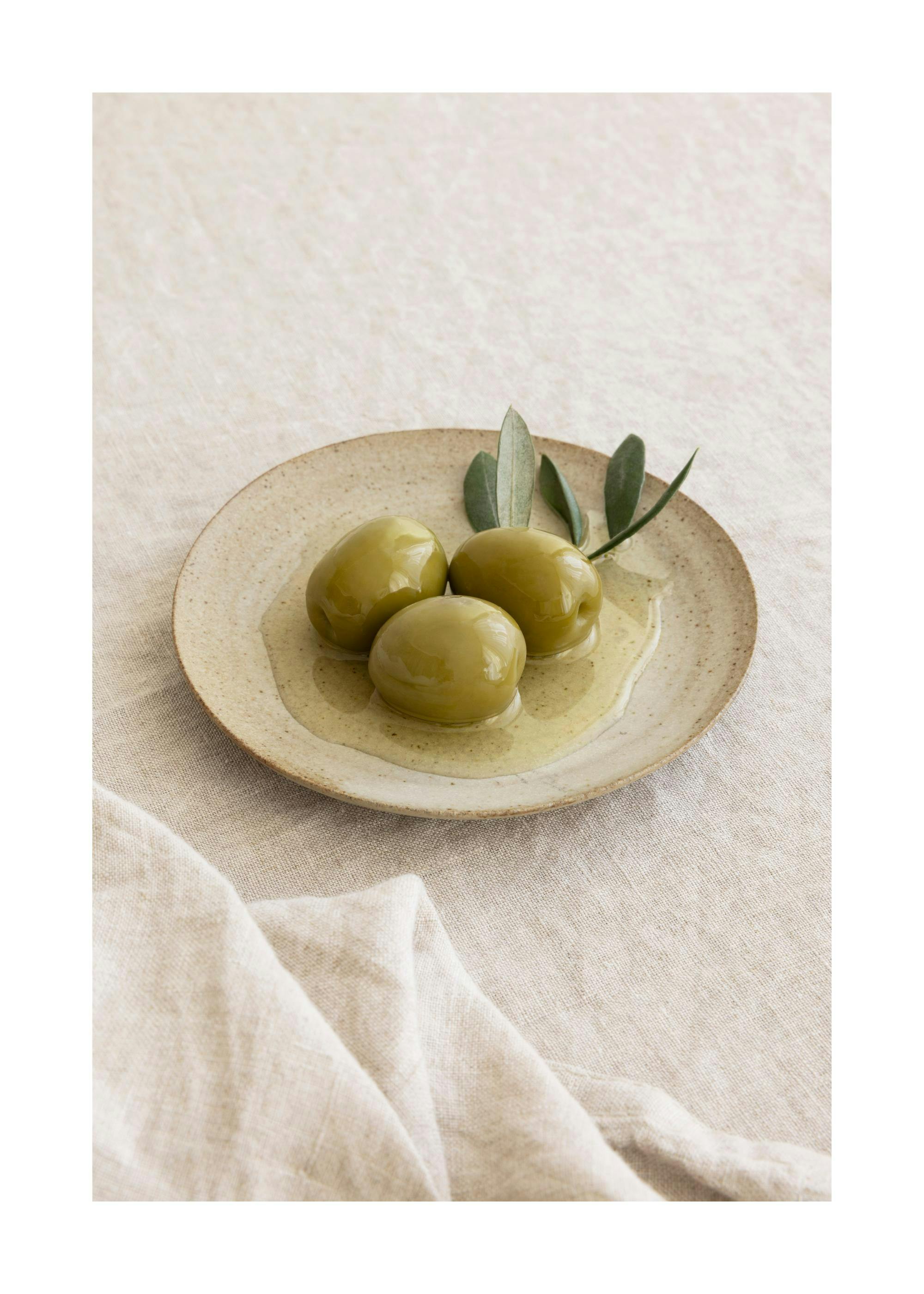 Drie groene olijven met een takje salie op een beige bord, met olijfolie op een linnen tafelkleed