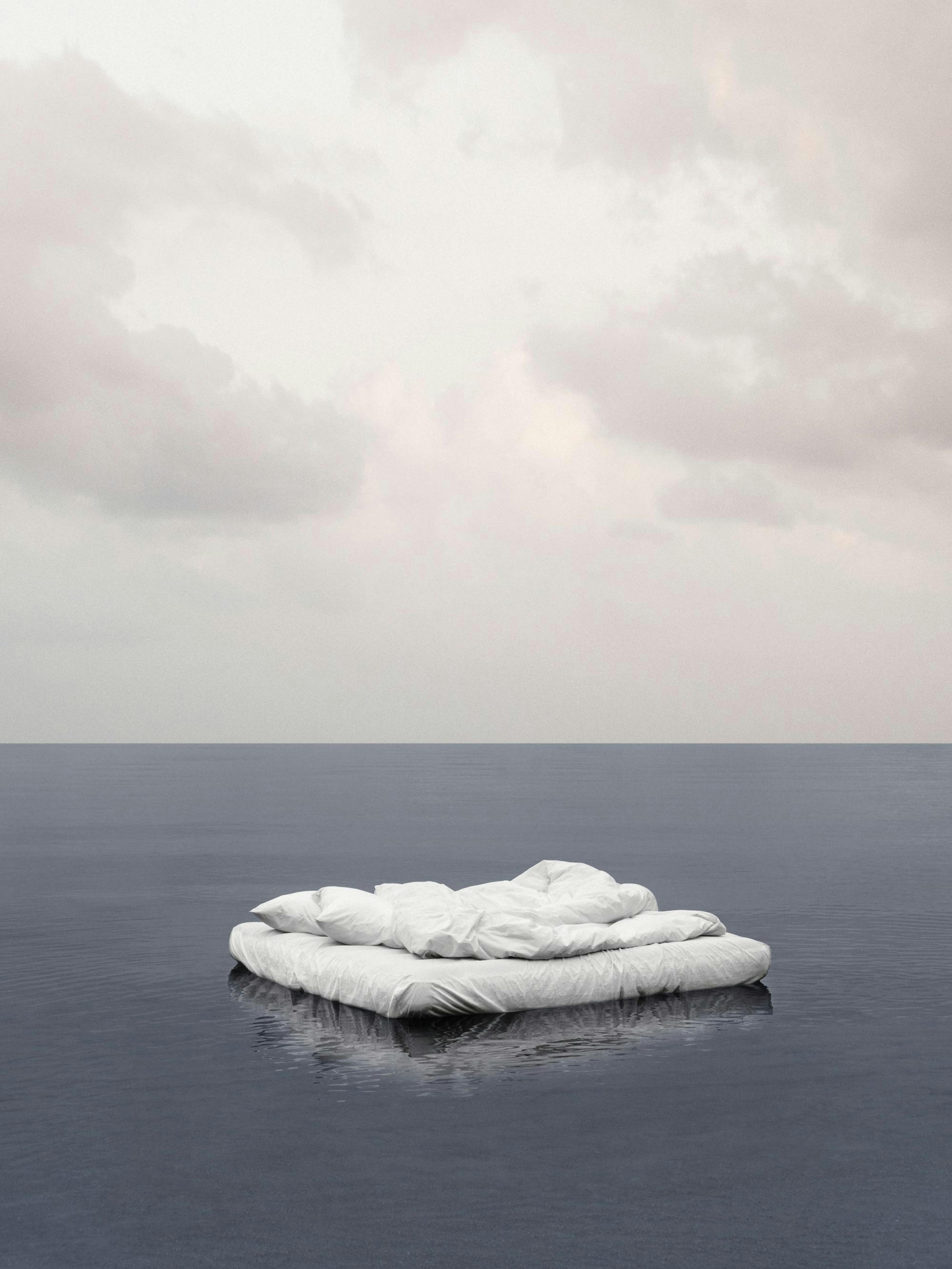 Póster fotográfico de una cama flotando en agua serena, con tonos grises y azules, textura granulada.