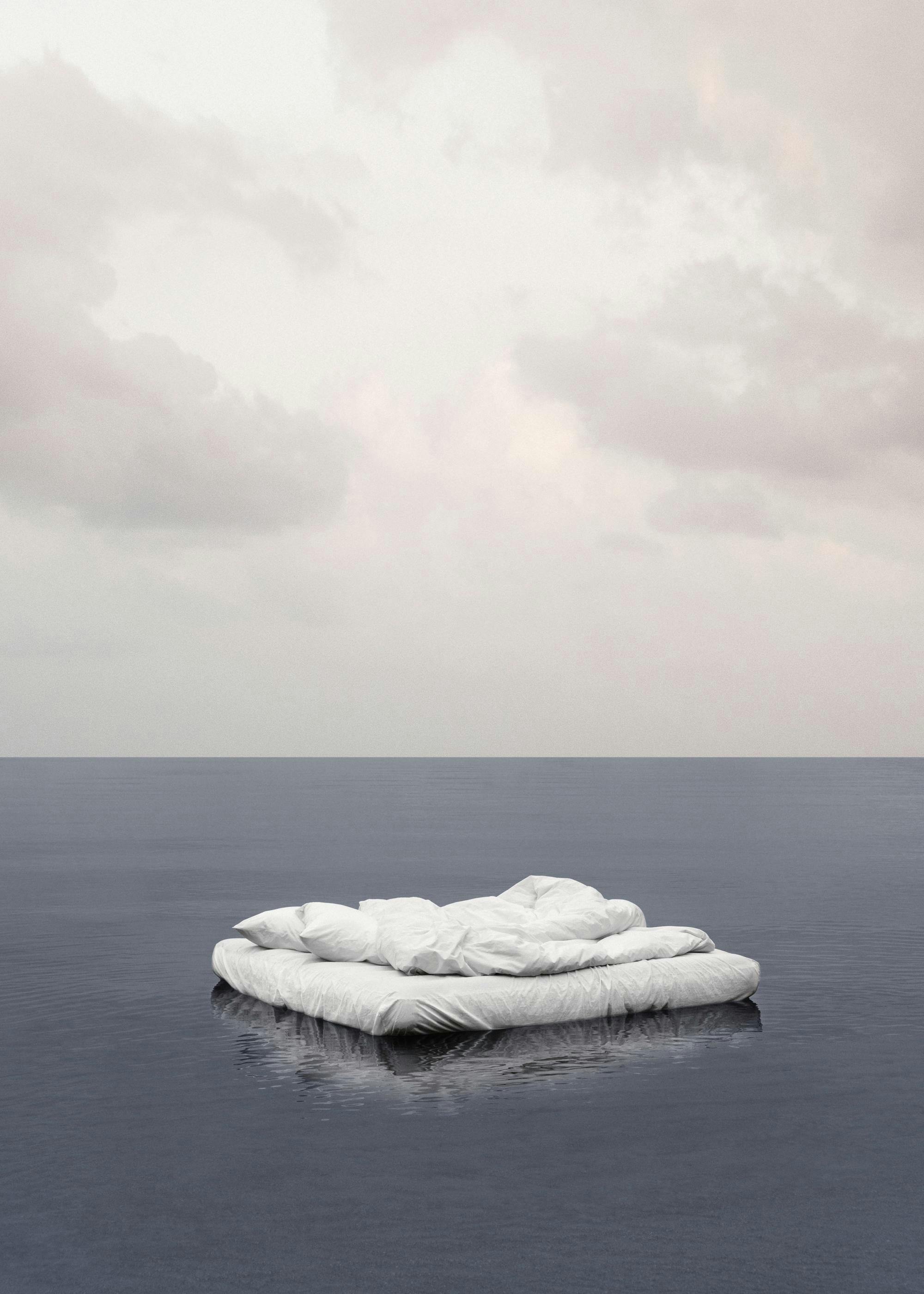 Dreamy photography print of a bed floating on water under a soft gray and blue sky, with a grainy texture.