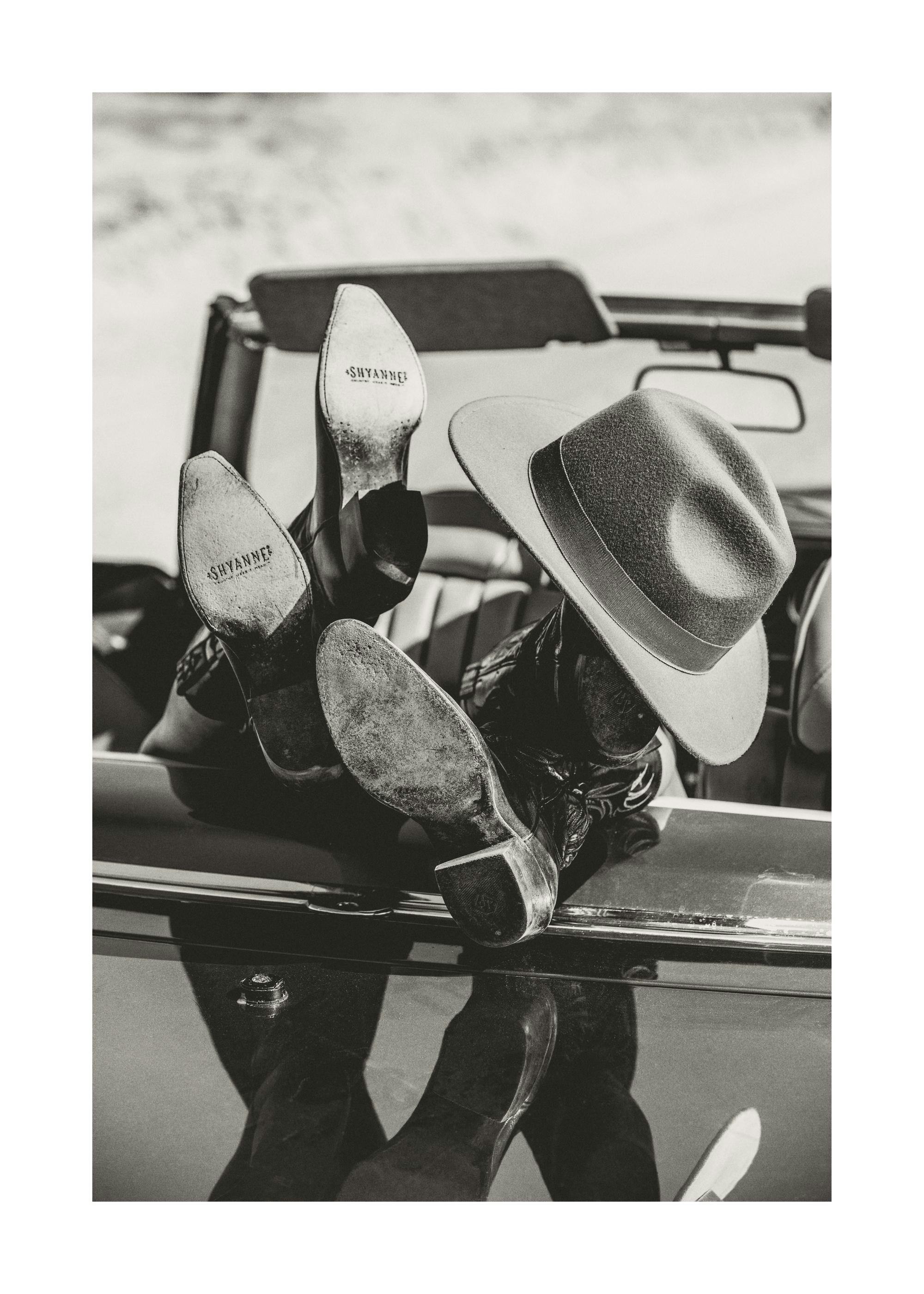 Black and white print of cowboy boots and hat resting on a car, perfect as a poster.
