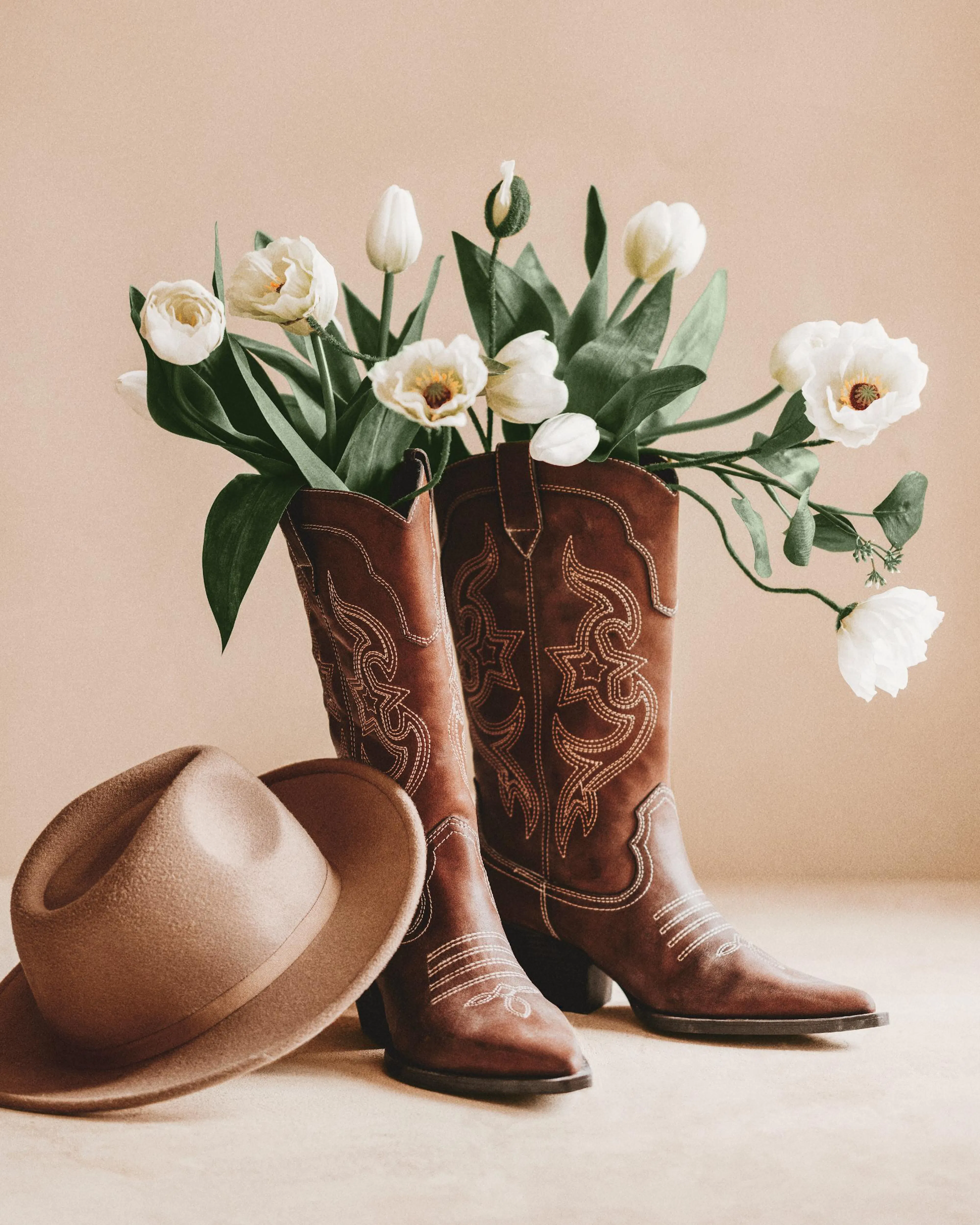 Ruskeat cowboy-saappaat täynnä valkoisia tulppaaneja ja pioneja, vieressään vaalea cowboy-hattu