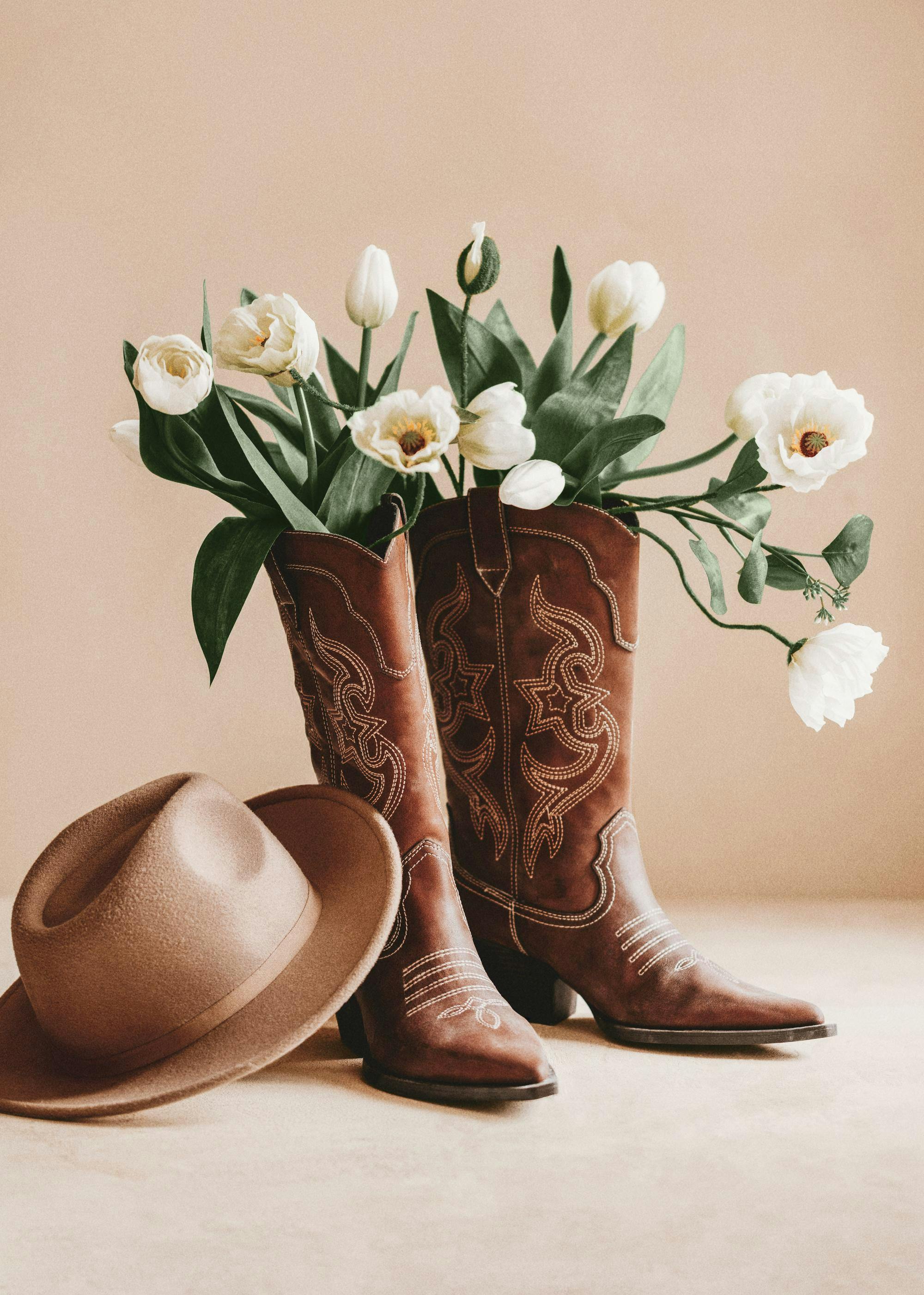 Braune Cowboystiefel mit floralen Stickereien, gefüllt mit weißen Tulpen und Pfingstrosen, daneben ein beiger Cowboyhut. Körnung