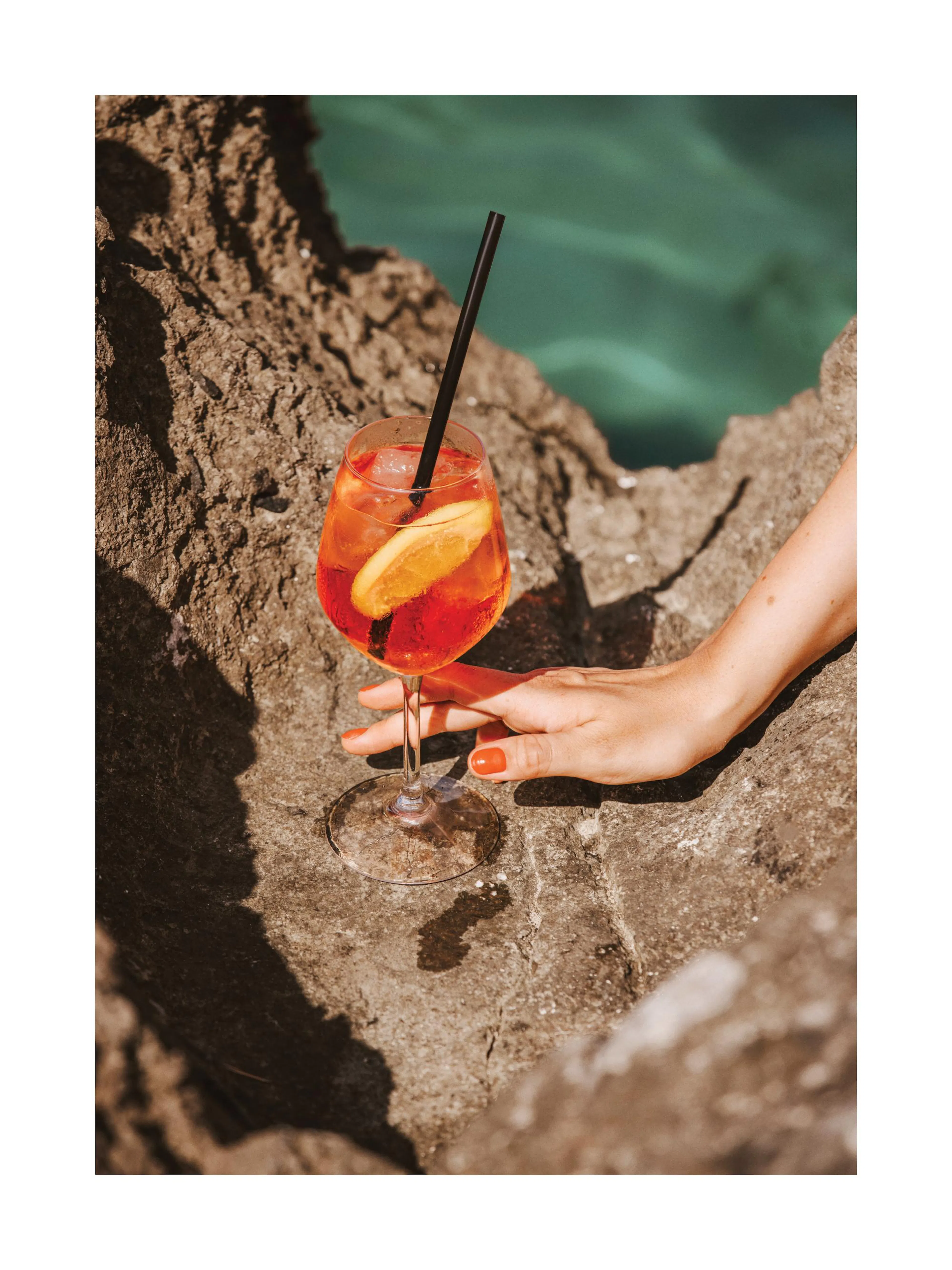 Spritz cocktail with orange slice on rocks by the ocean, hand with orange nails resting on rock. Holiday drink poster.