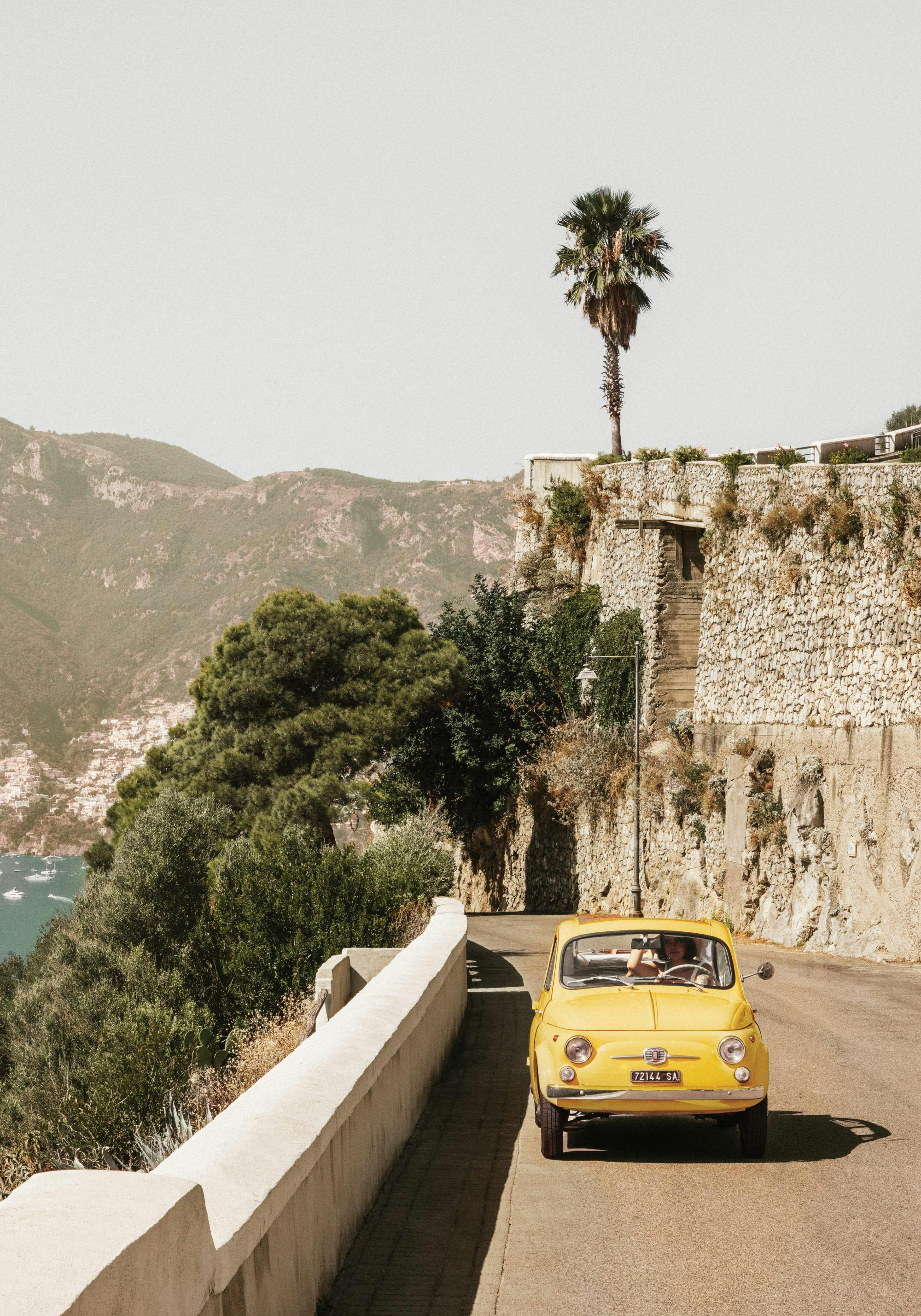 Gul bil på Amalfikusten, Italien. Kustväg med palmer, berg och hav i bakgrunden.