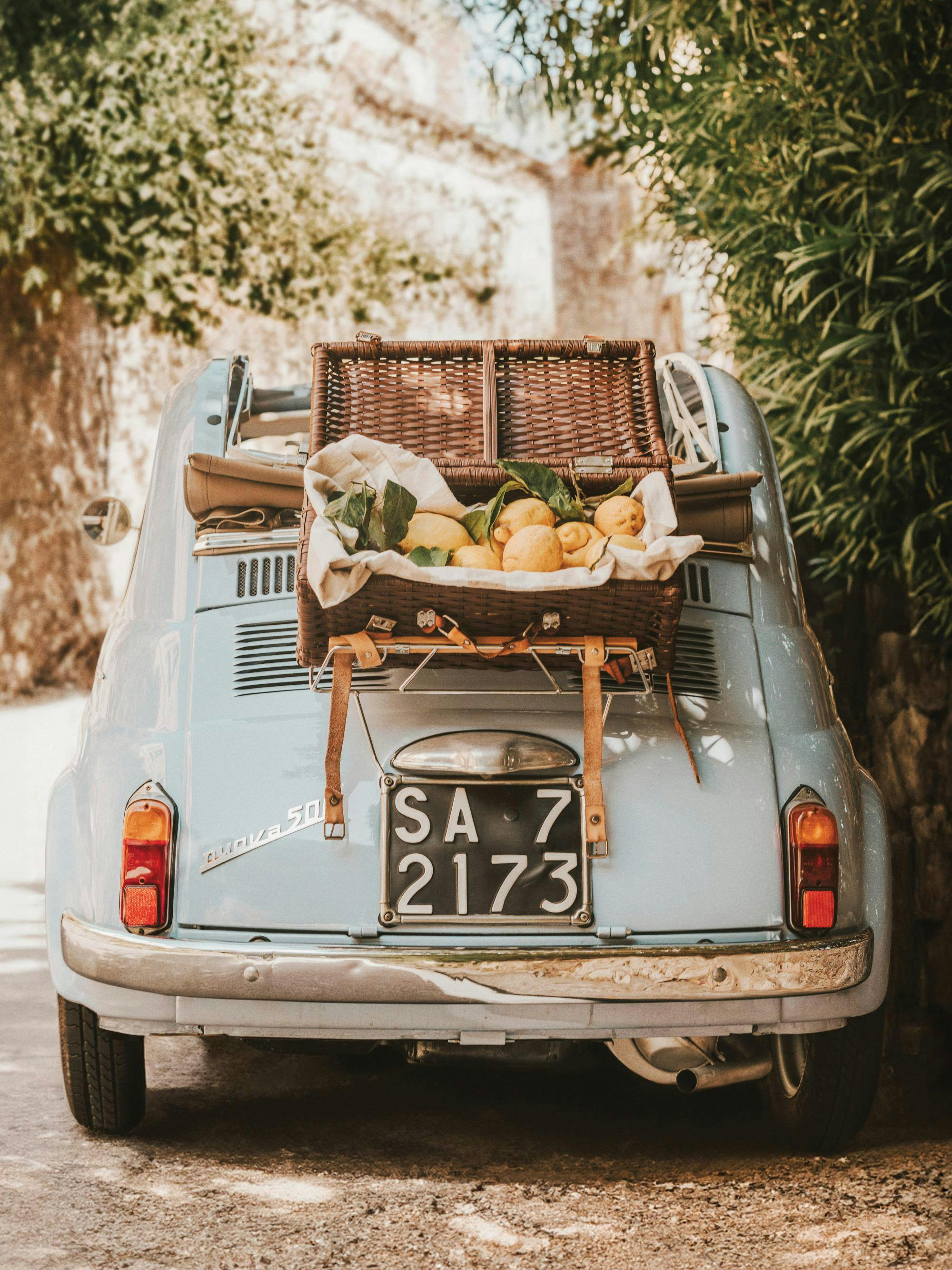 Fotografi av en ljusblå vintagebil med en korg fylld med citroner baktill, omgiven av grönska.
