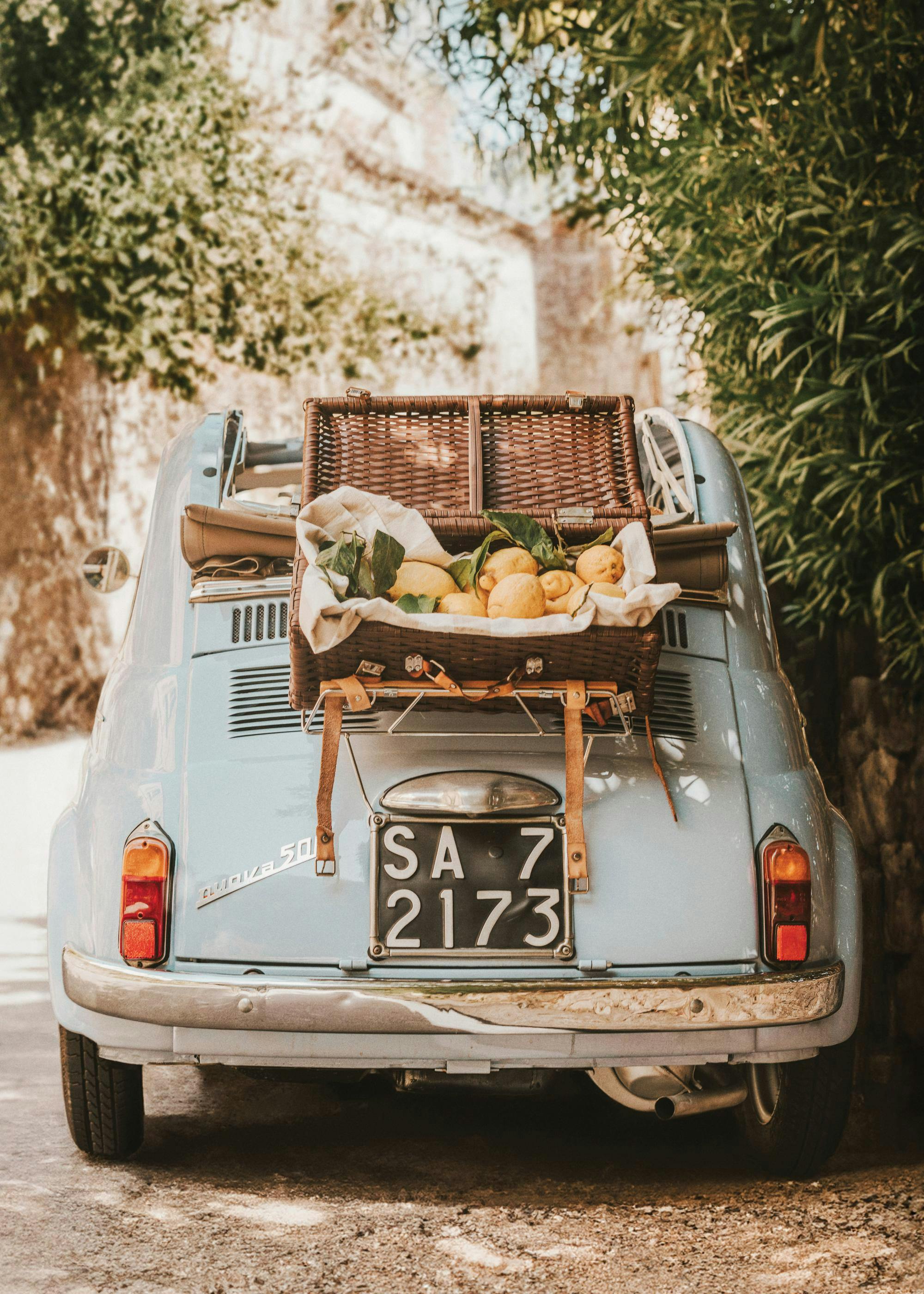 Fotografi av en ljusblå vintagebil med en korg fylld med citroner baktill, omgiven av grönska.