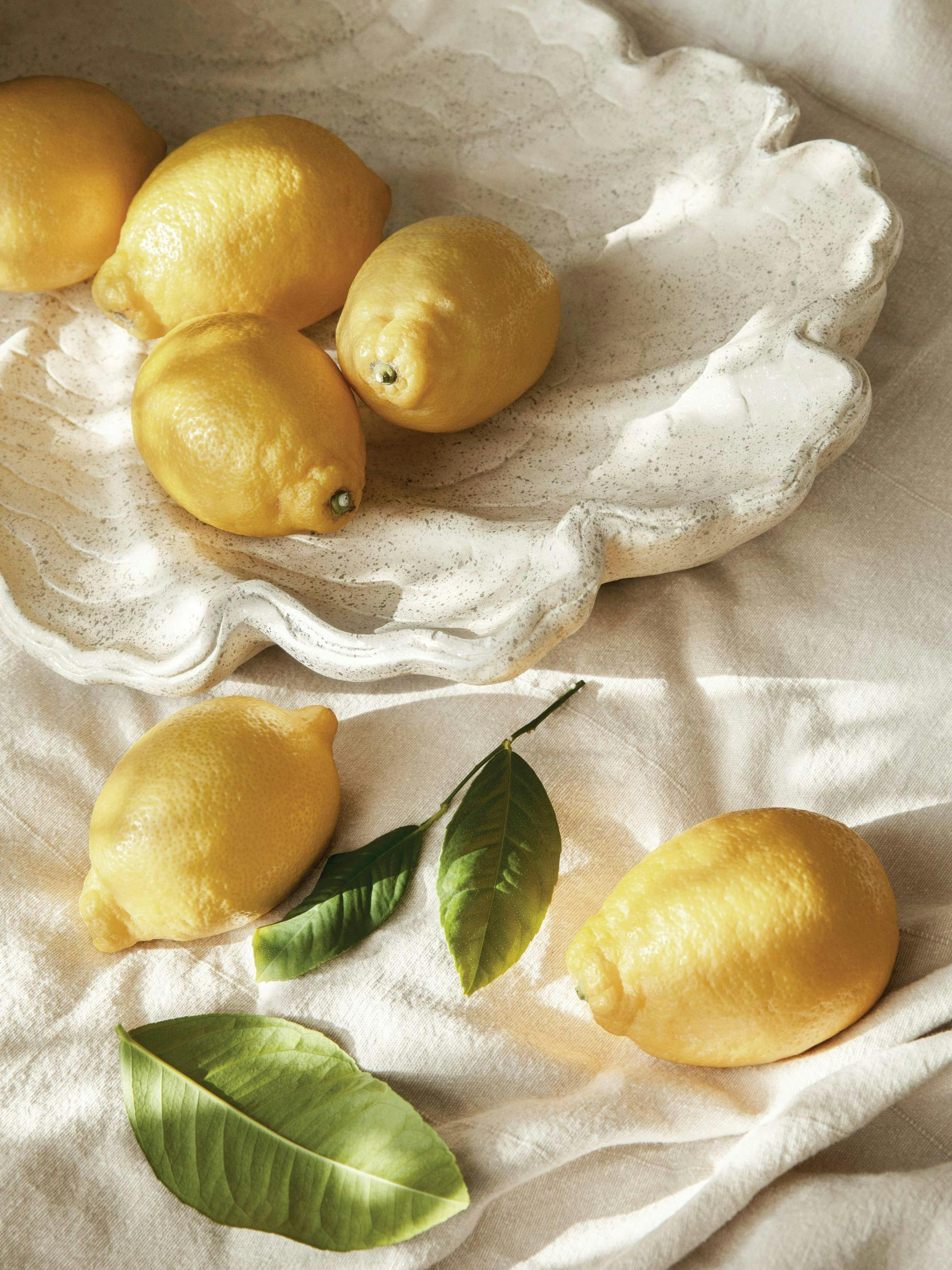 Gula citroner och gröna blad på en vit vågig skål och en texturerad vit duk, somrig köksinredning.