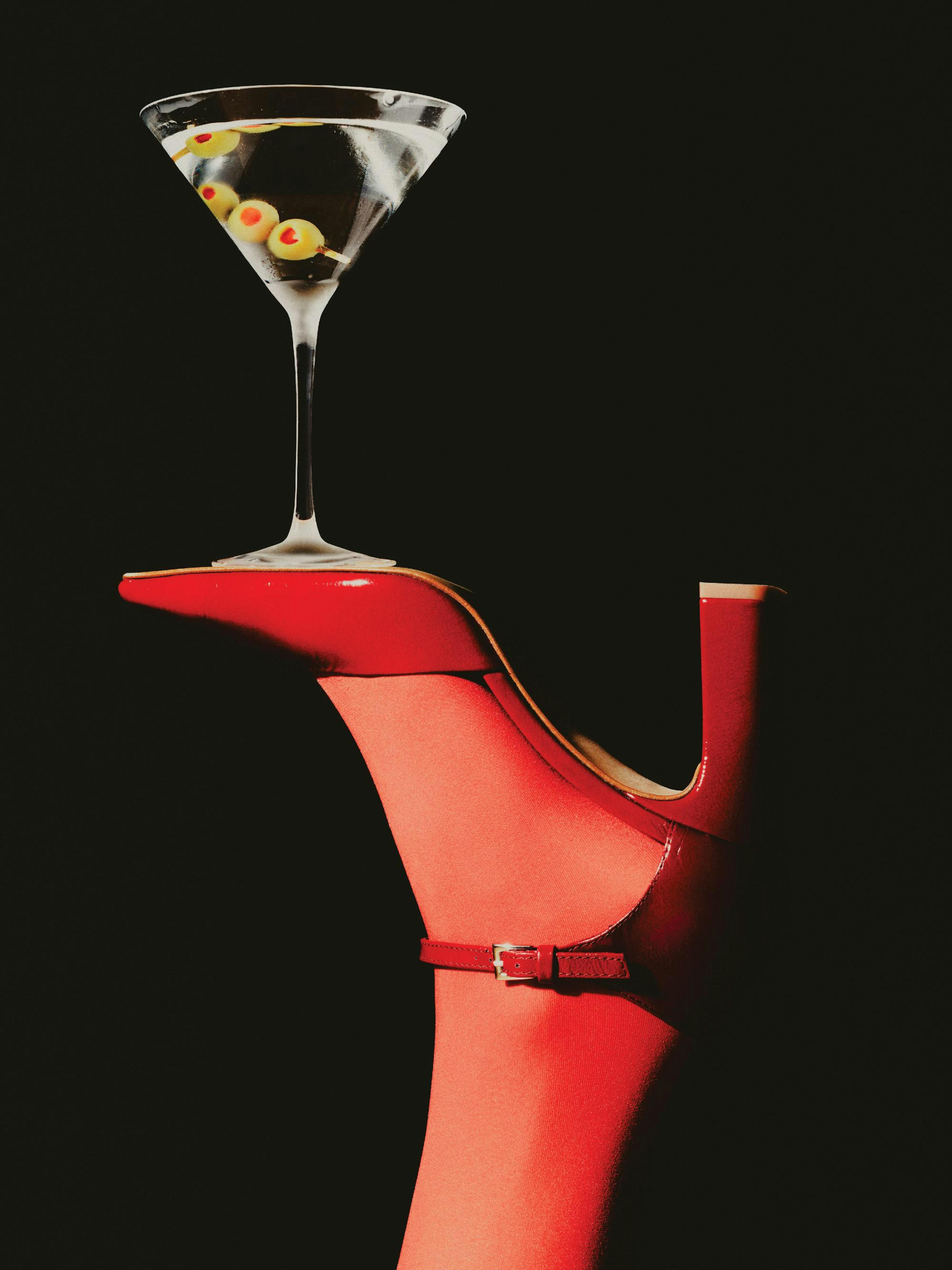 Plakát s koktejlem martini balancujícím na špičce červené lodičky, elegantní černobílá fotografie s červenými detaily.