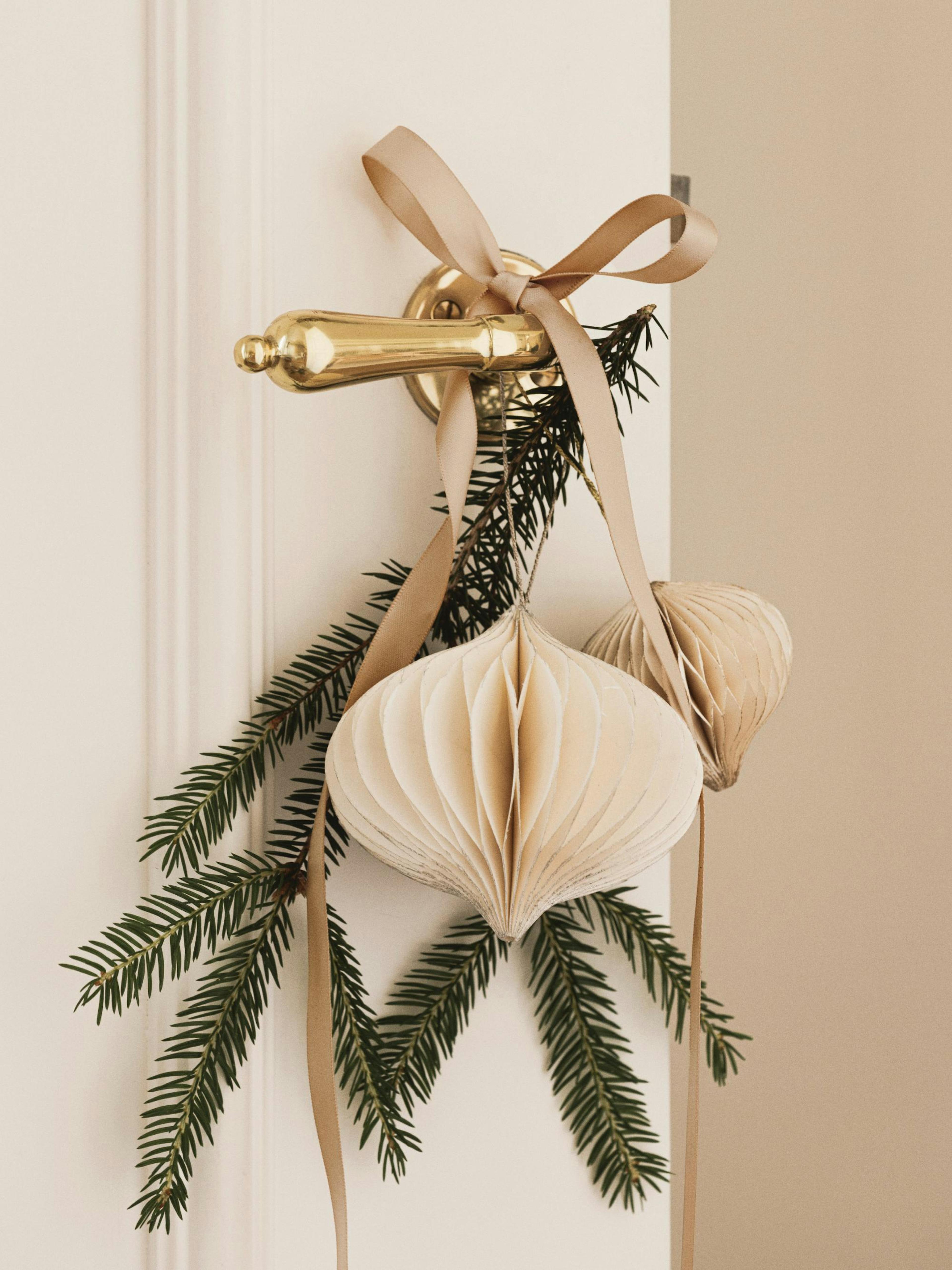 Decorazione natalizia per porta con rami di pino, nastro beige e ornamenti in carta.