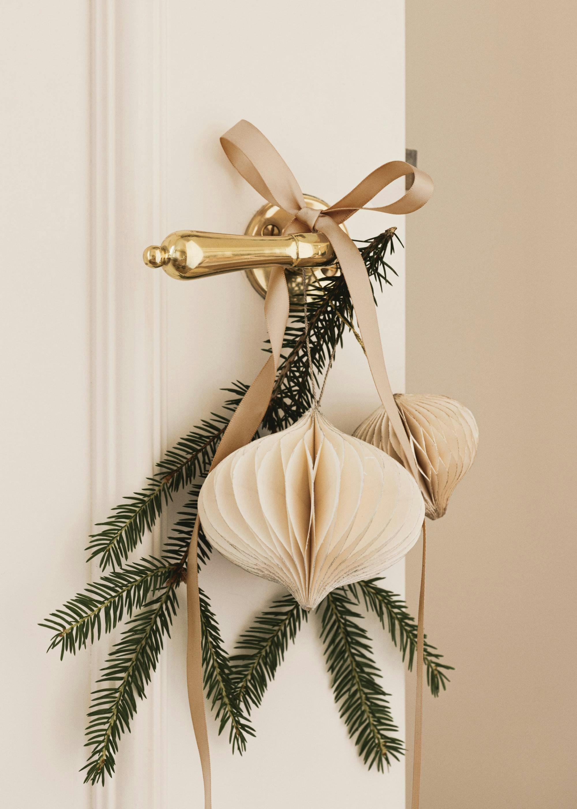 Decorazione natalizia per porta con rami di pino, nastro beige e ornamenti in carta.