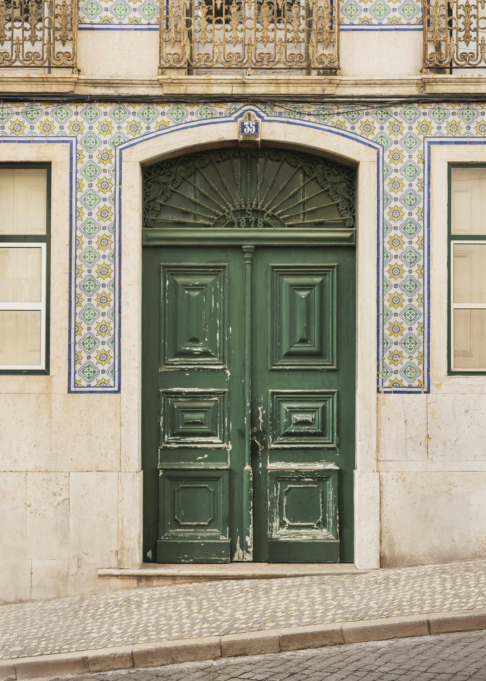 A print of a weathered green door with a decorative transom, flanked by yellow, green, and blue tile work on a beige building.